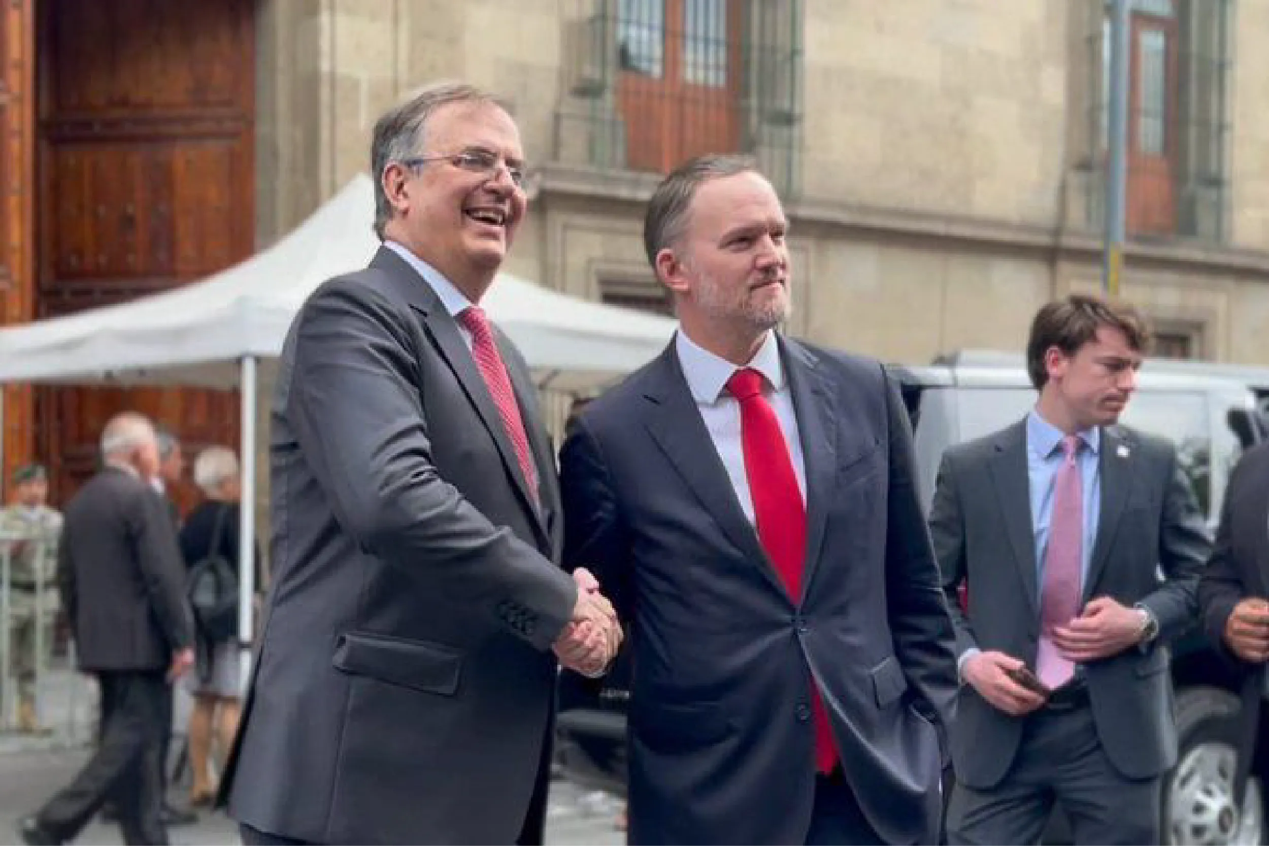 Two businessmen in suits shaking hands outdoors at a formal event, smiling in the street.
