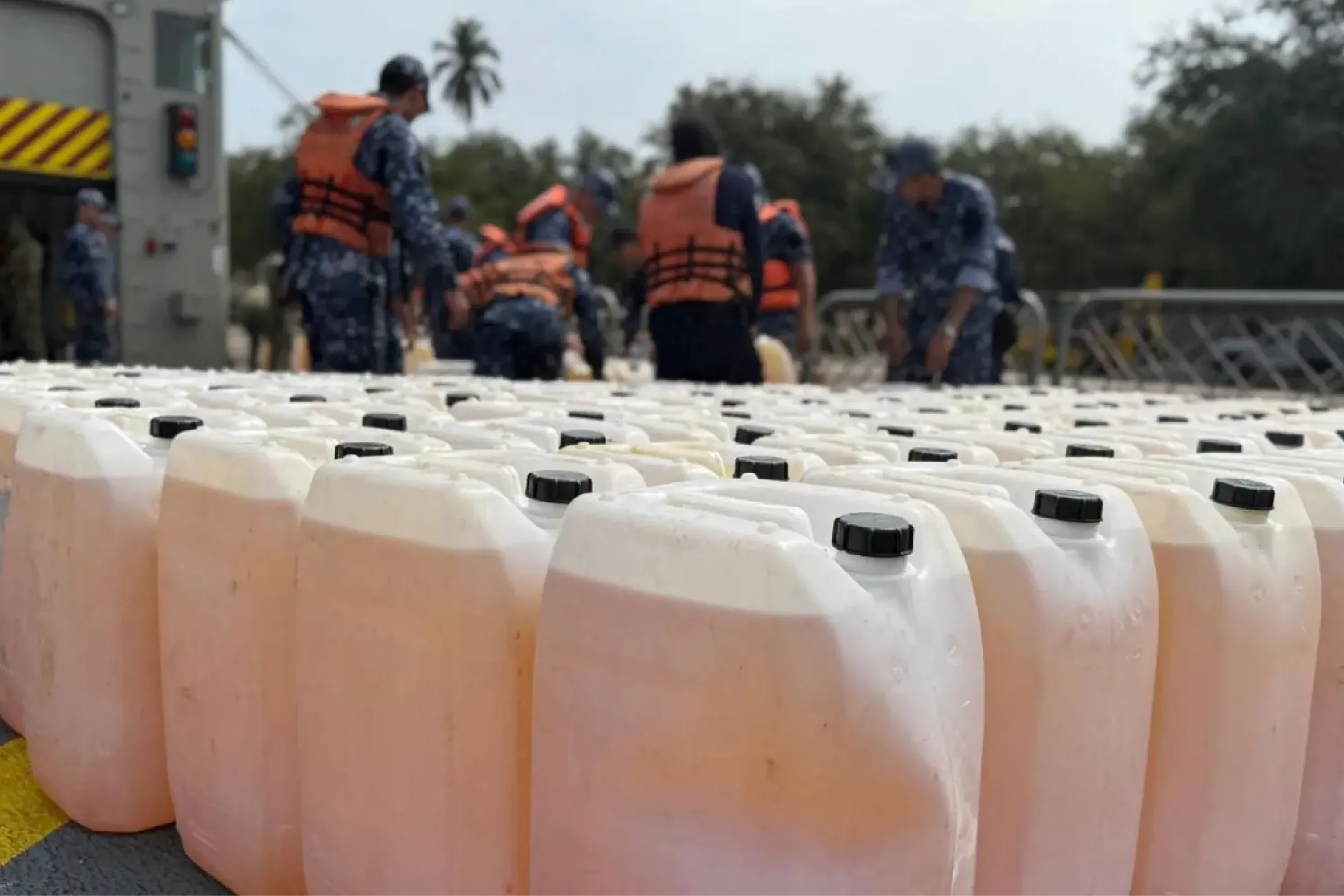 Row of white plastic jerry cans with black caps in the foreground, while people wearing orange life jackets stand on a dock in the background.
