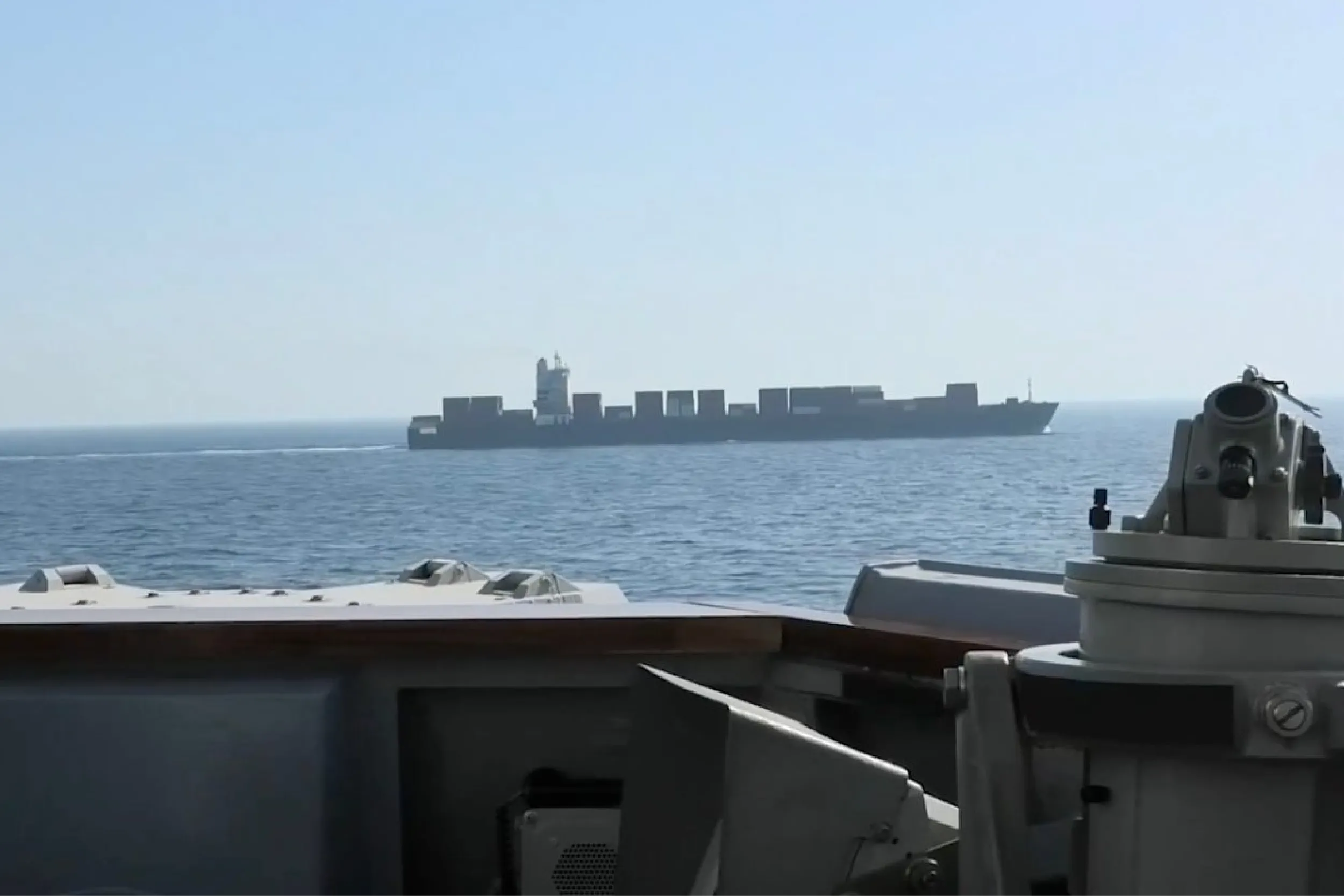 Container ship on calm sea seen from a navy vessel’s deck, with a gun turret in the foreground on the right.