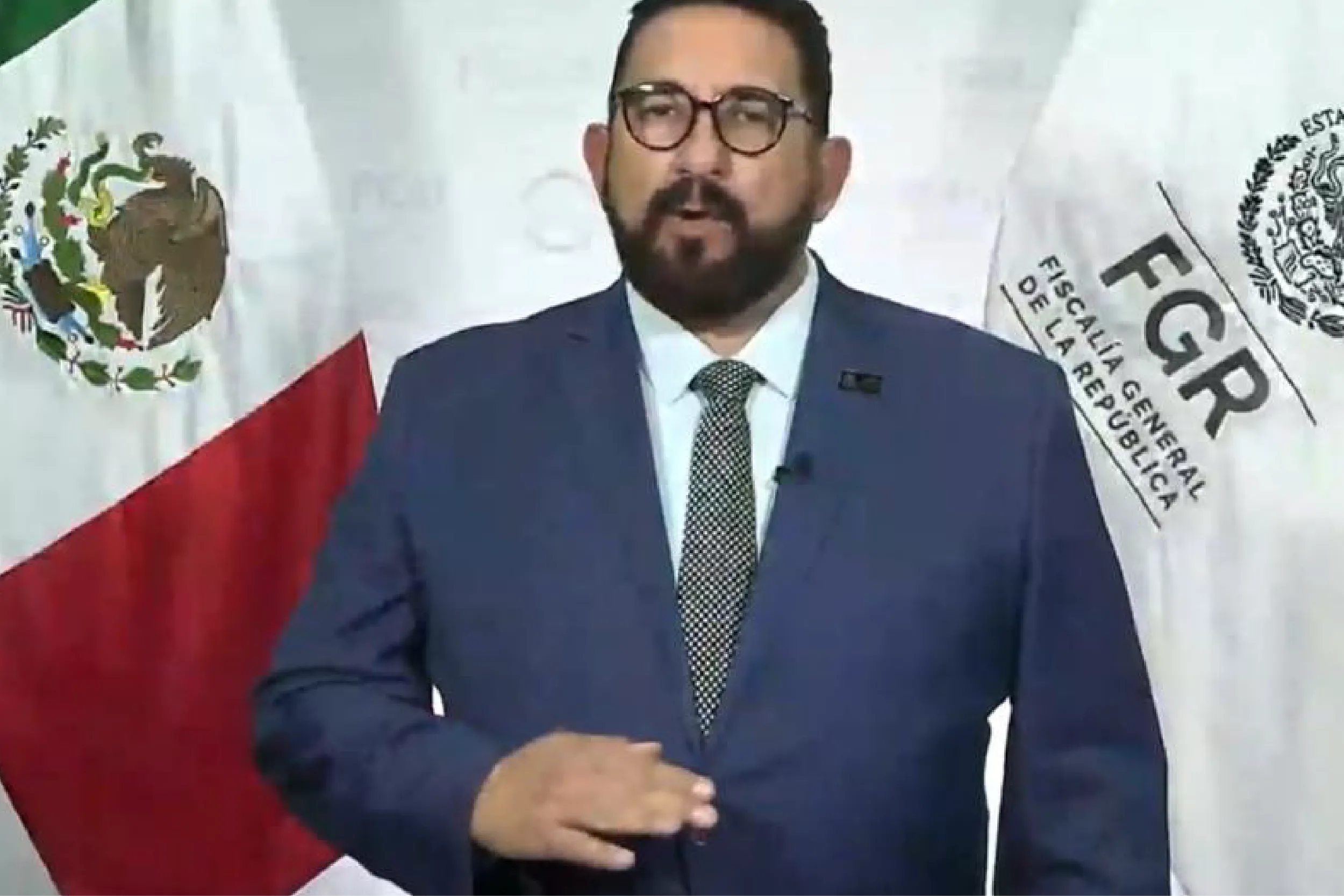 Man in a dark suit and dotted tie speaks at a formal event, with national flags in the background and a beard and glasses.