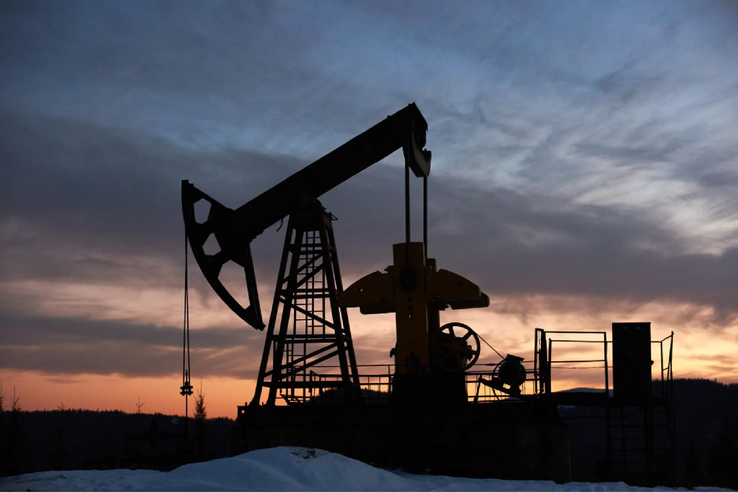 Silhouette of an oil pumpjack against a colorful sunset sky, with snow on the ground in the foreground.