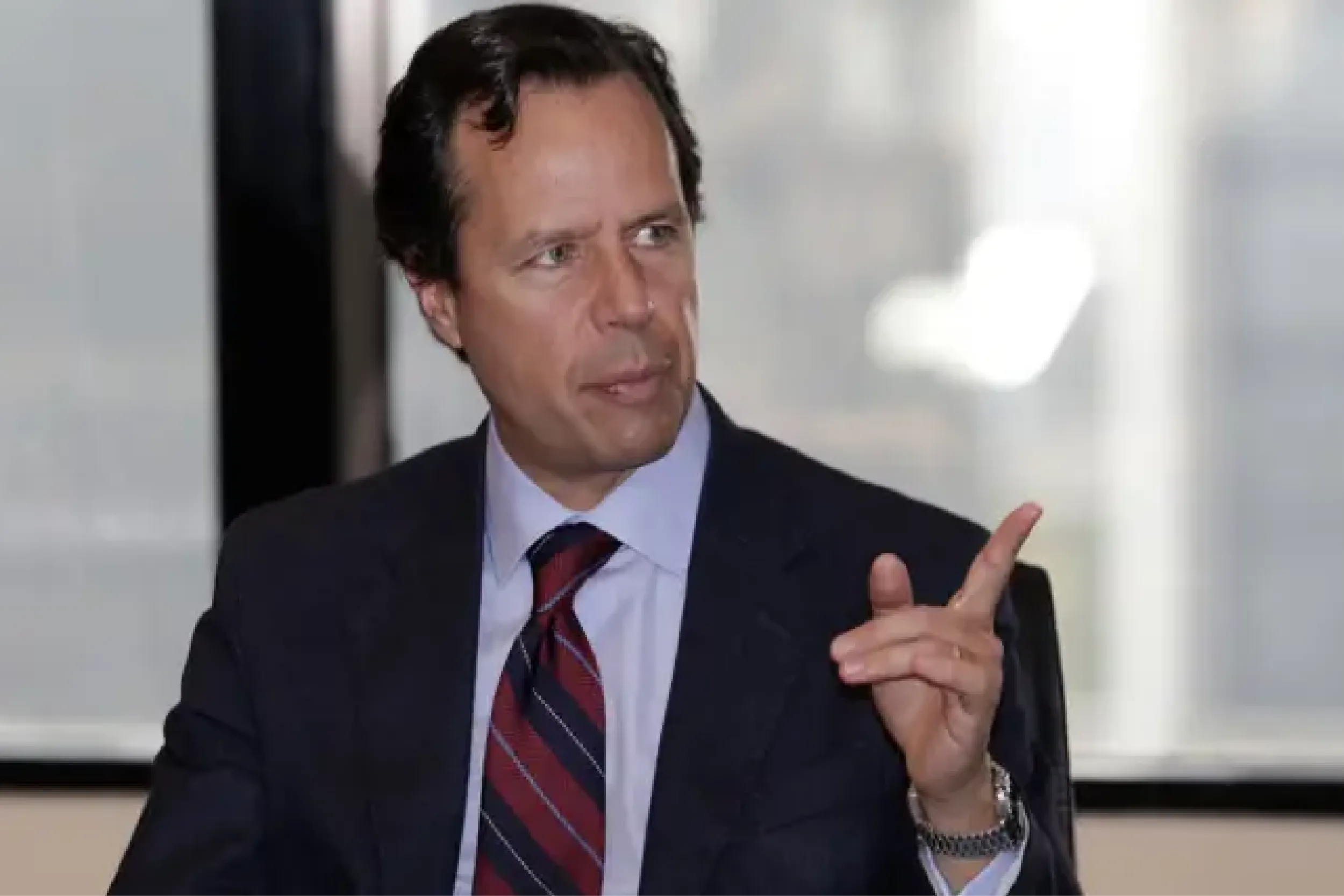 Man in a dark suit and striped tie gestures with his hands while speaking in a professional setting.