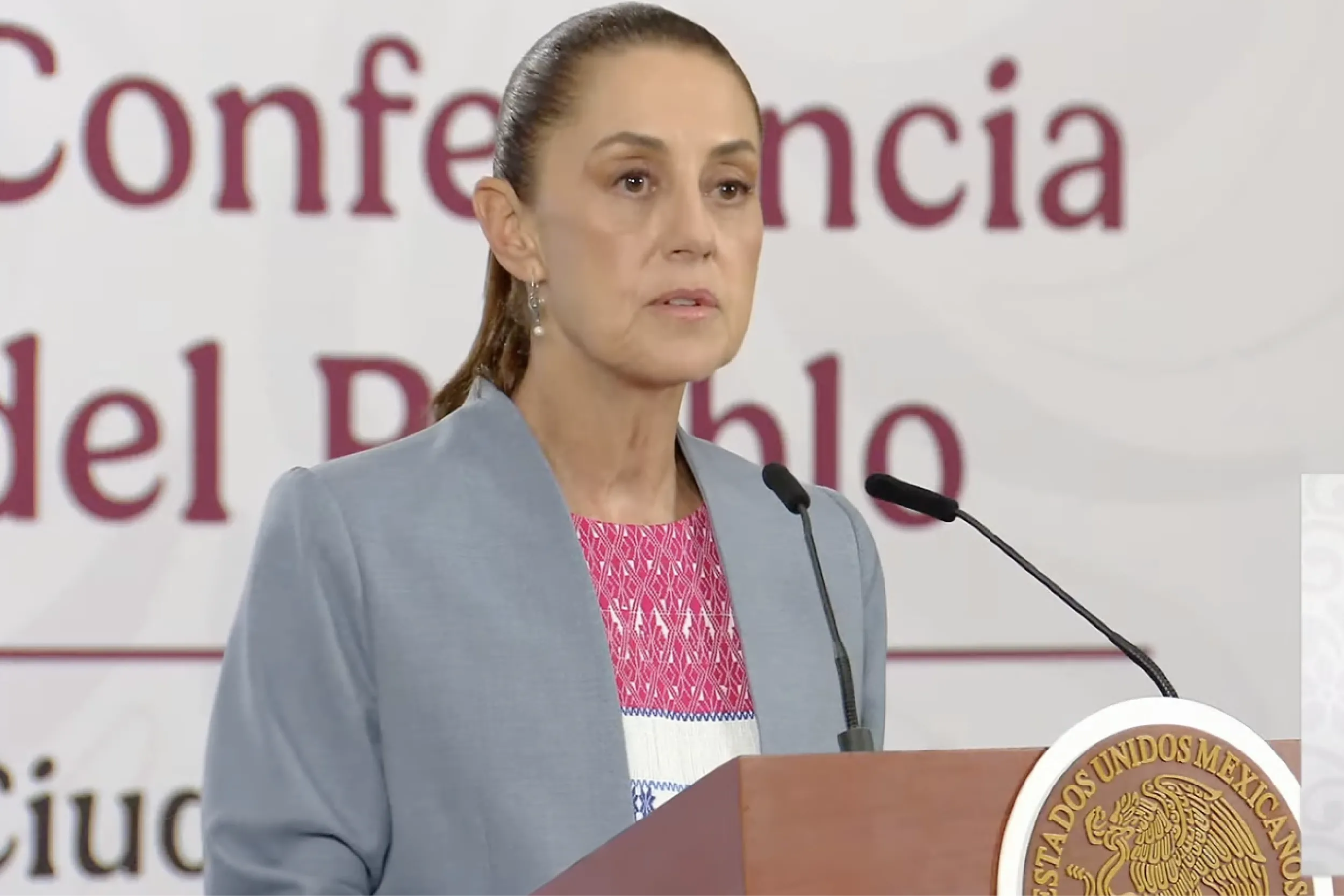Female official in a blue blazer delivering a speech at a podium with the Mexican seal and microphones visible at a press conference background text behind her