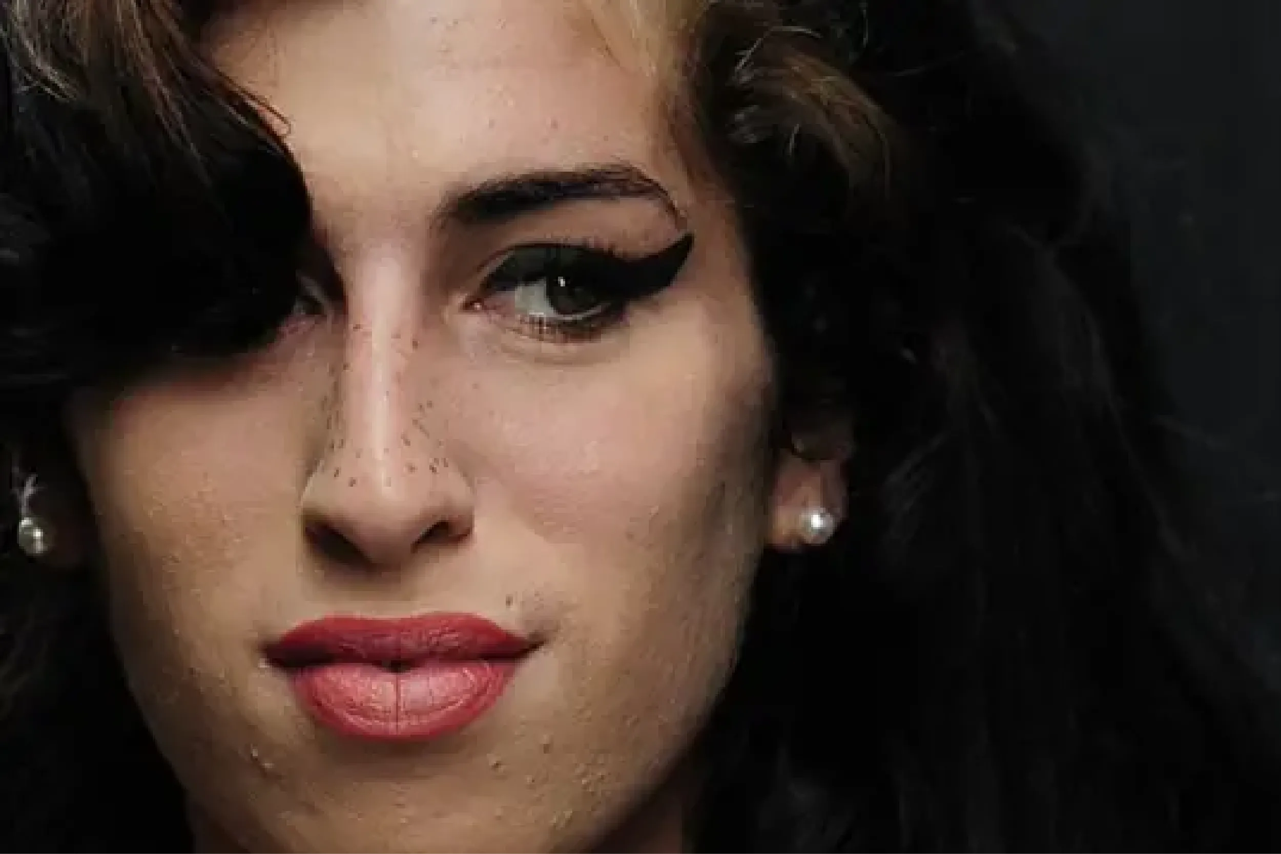 Close-up of a woman's face showing winged eyeliner, red lips, and pearl earrings with dark curly hair nearby.