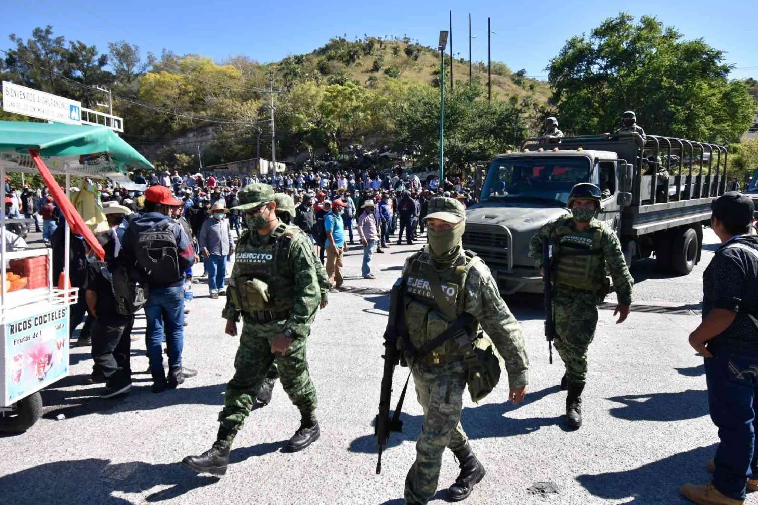 Soldiers in camouflage with rifles walk through a crowded street beside a large military truck on a sunny day.
