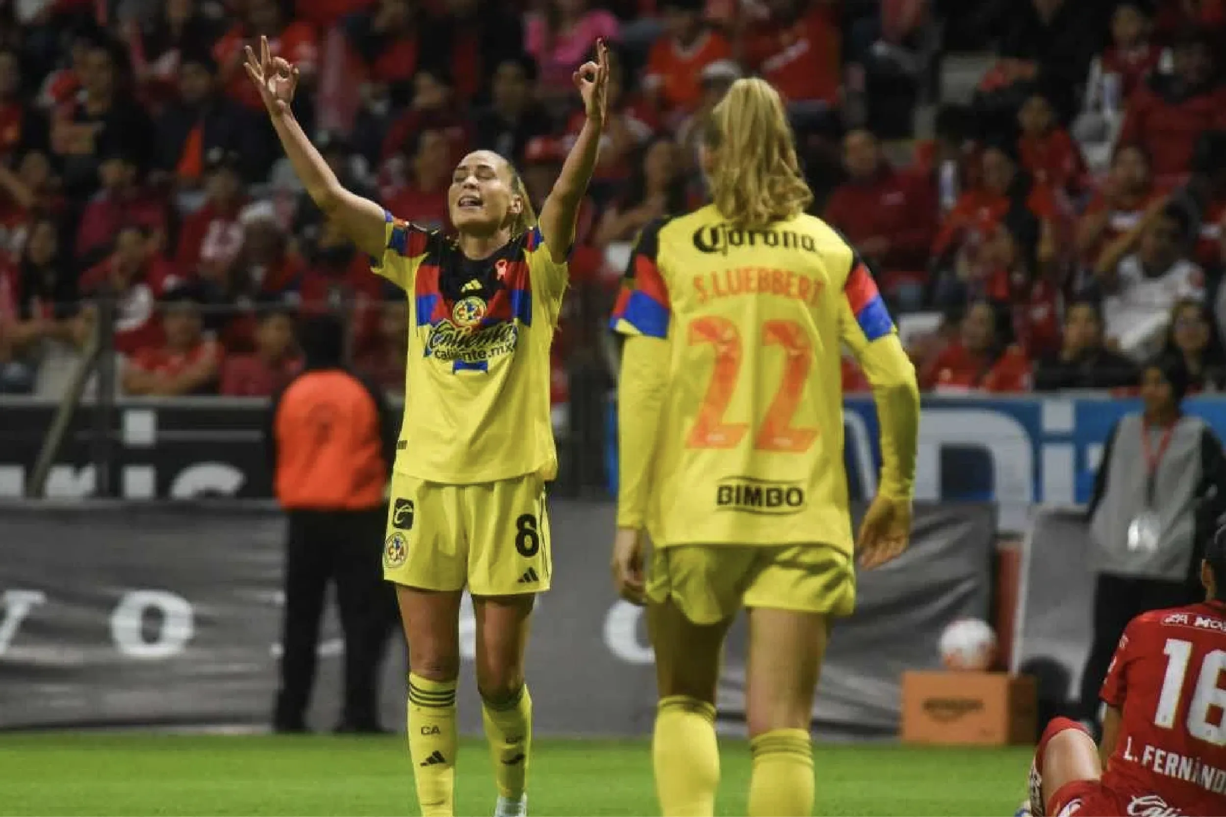 Two soccer players in yellow uniforms celebrate on the field with arms raised, crowd cheering in the stands behind them.
