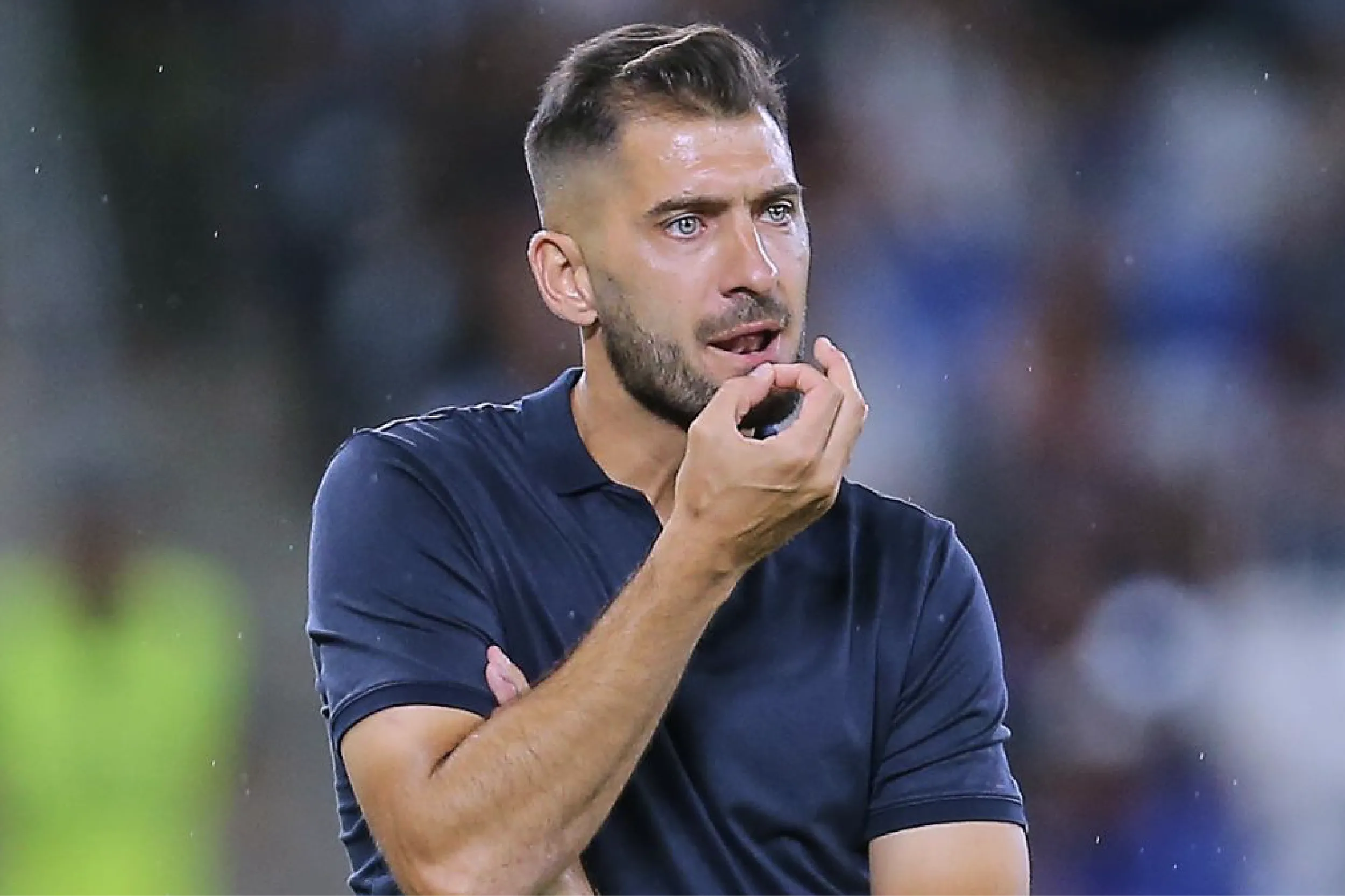 Man standing outdoors in a navy polo, hand near mouth in a thoughtful pose, looking ahead.