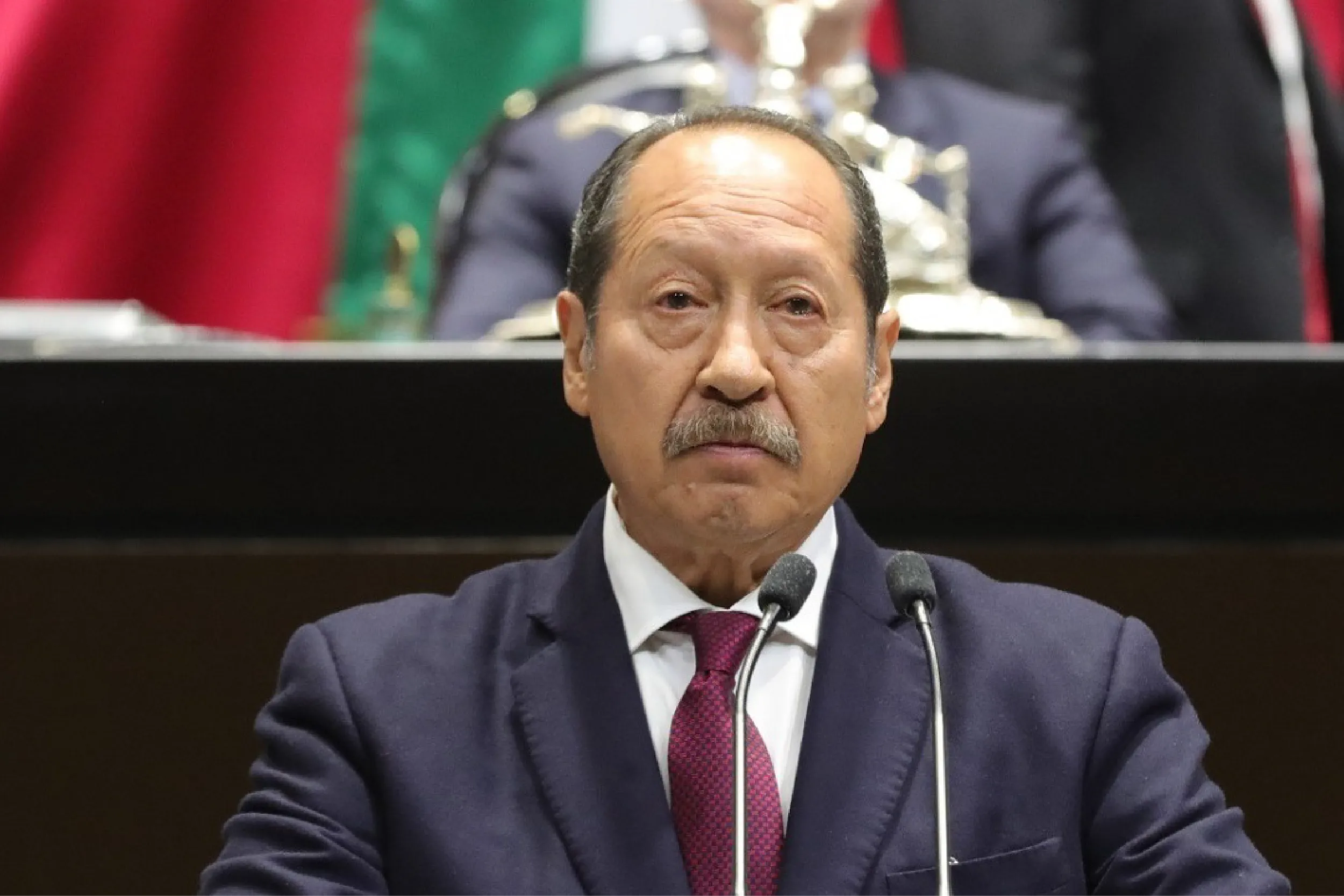 A middle-aged man in a navy suit and red tie speaks at a podium with two microphones. Behind him are blurred flags and official decor.