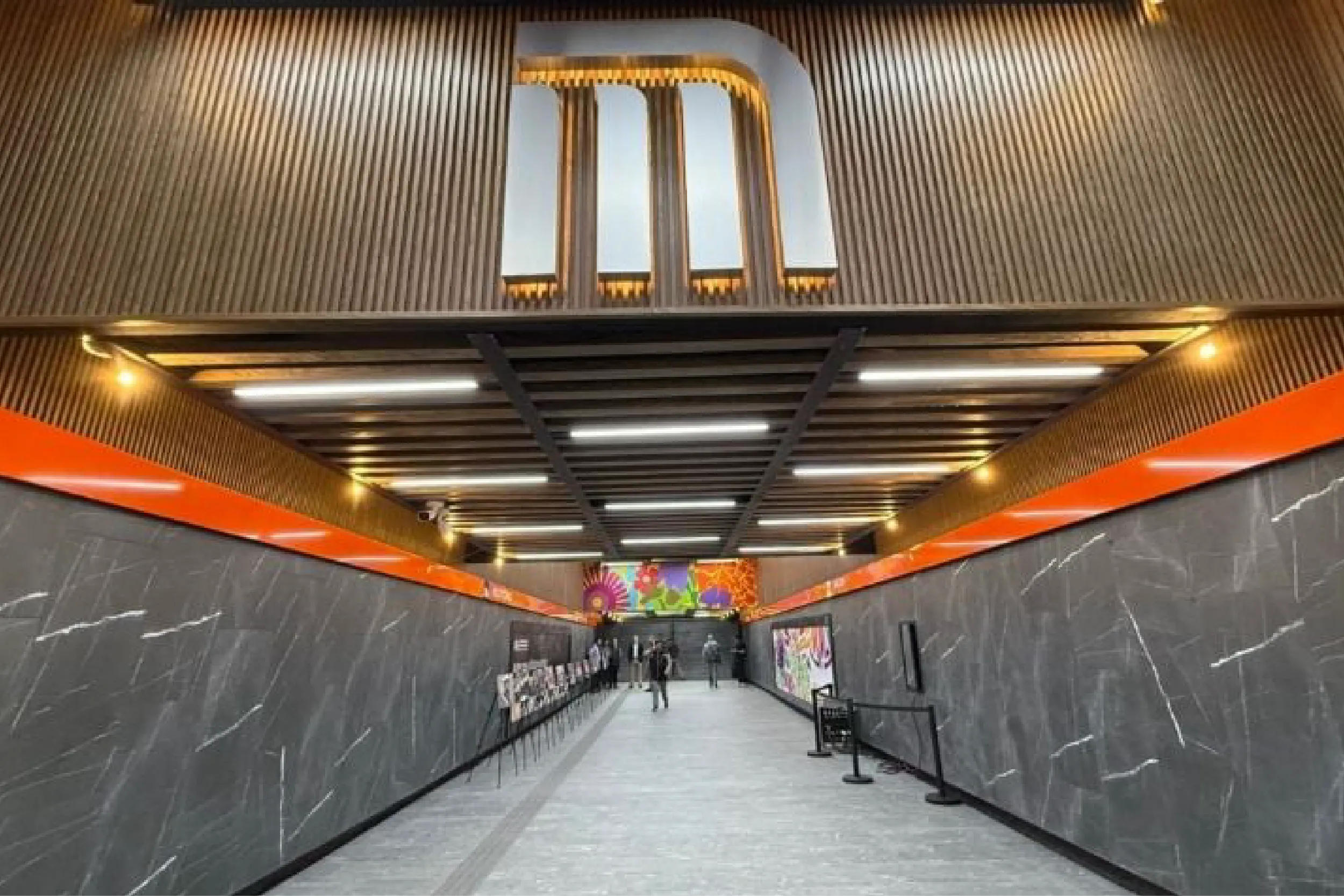 Long underground subway corridor with dark marble walls, orange accents, and bright ceiling lights; a few passengers walk toward a colorful mural in the distance.