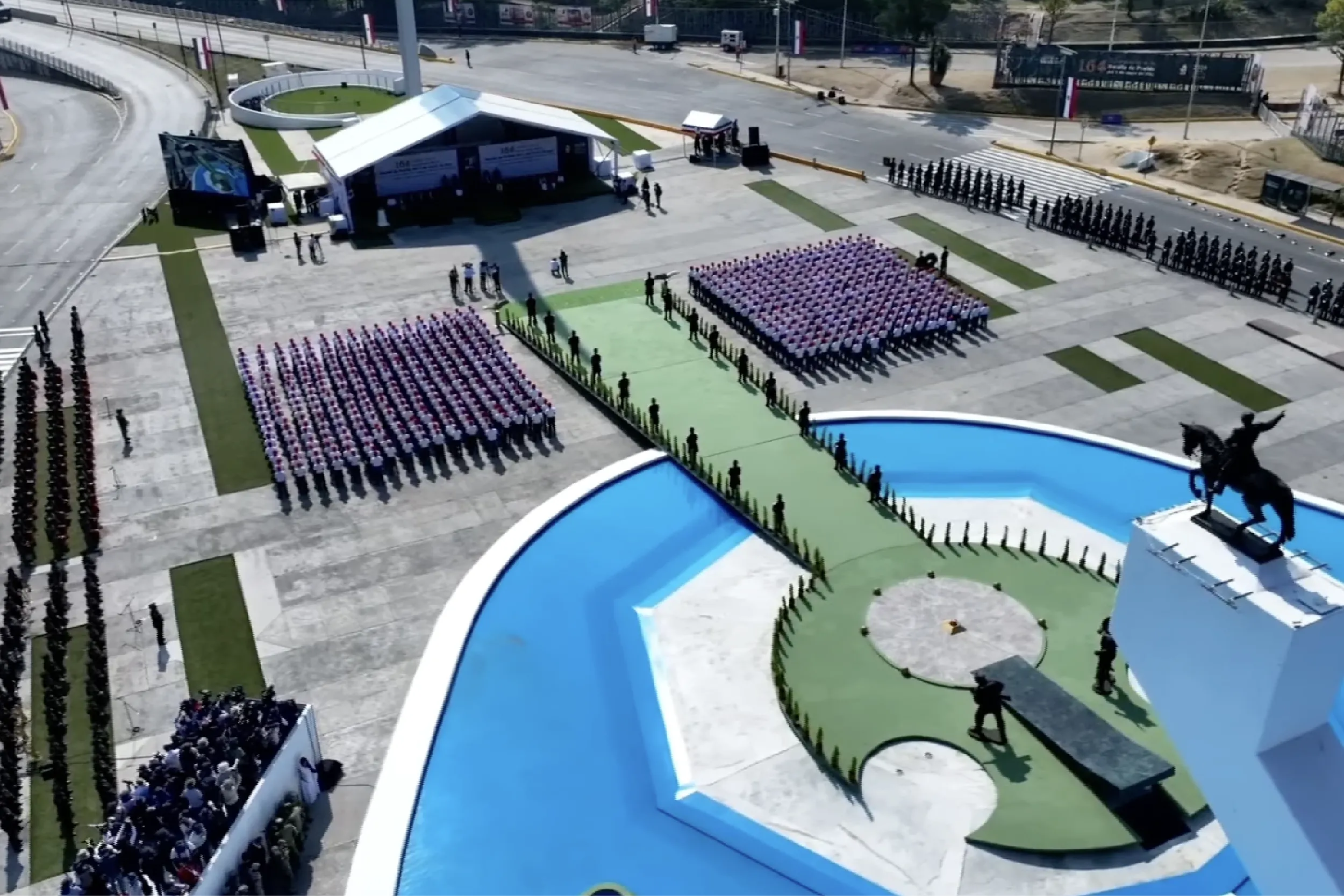 Aerial view of a military ceremony in a sunlit plaza with rows of uniformed soldiers standing at attention around a white tent stage and spectators along the edges