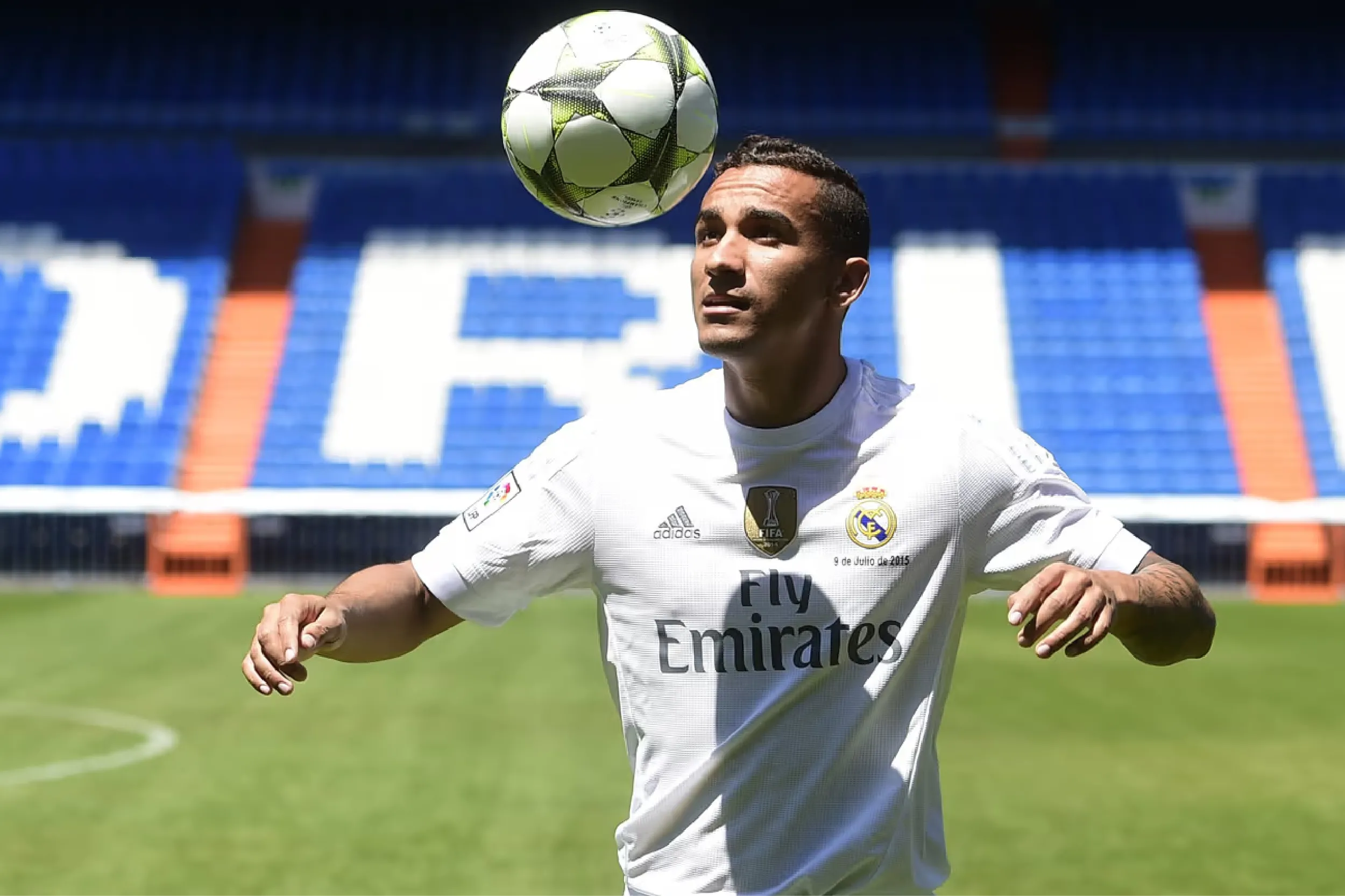 Soccer player in a white Real Madrid jersey heads a ball in a sunny stadium, with blue seats in the background.