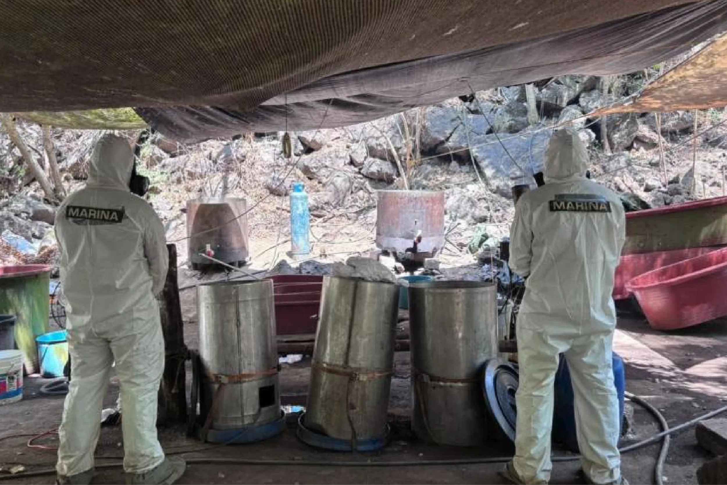 Two workers in white protective suits labeled MARINA stand under a shelter, inspecting large metal drums in a rocky outdoor workspace.