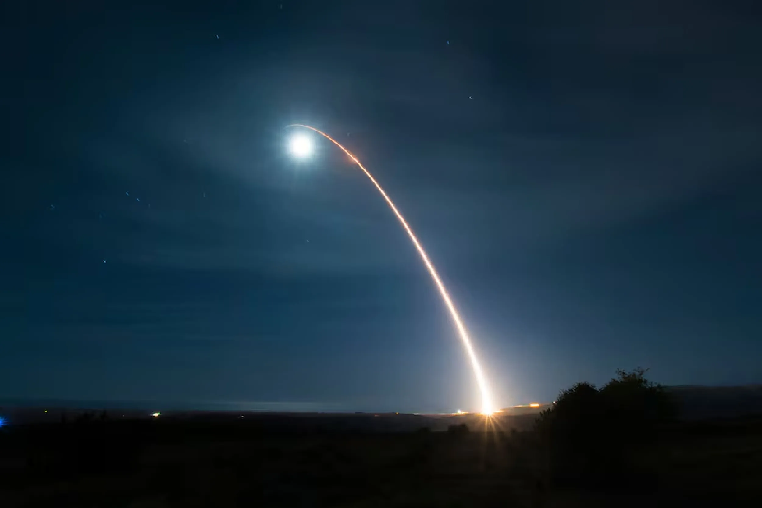Nighttime rocket launch with a bright plume and an orange curved exhaust trail across the dark sky, over a low landscape.