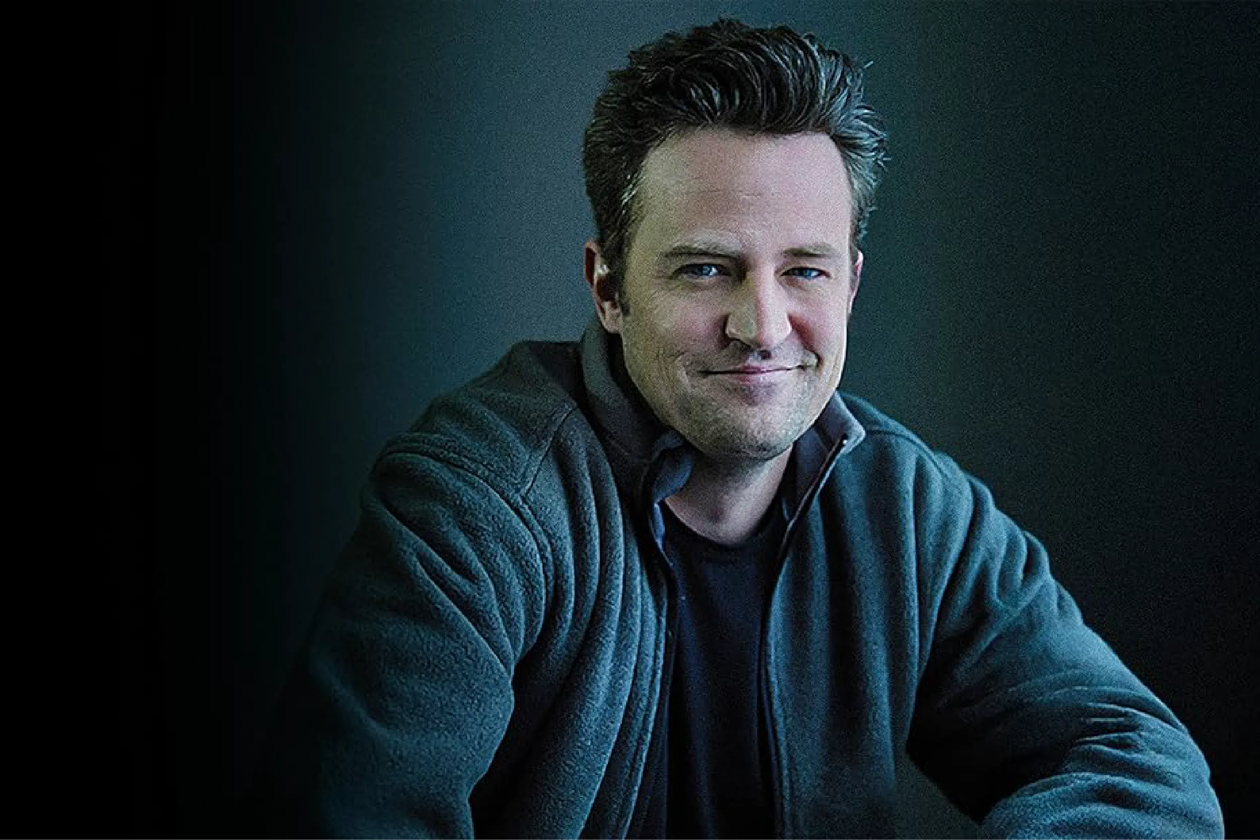 Portrait of a smiling man with dark hair in a blue sweater against a dark gradient background.