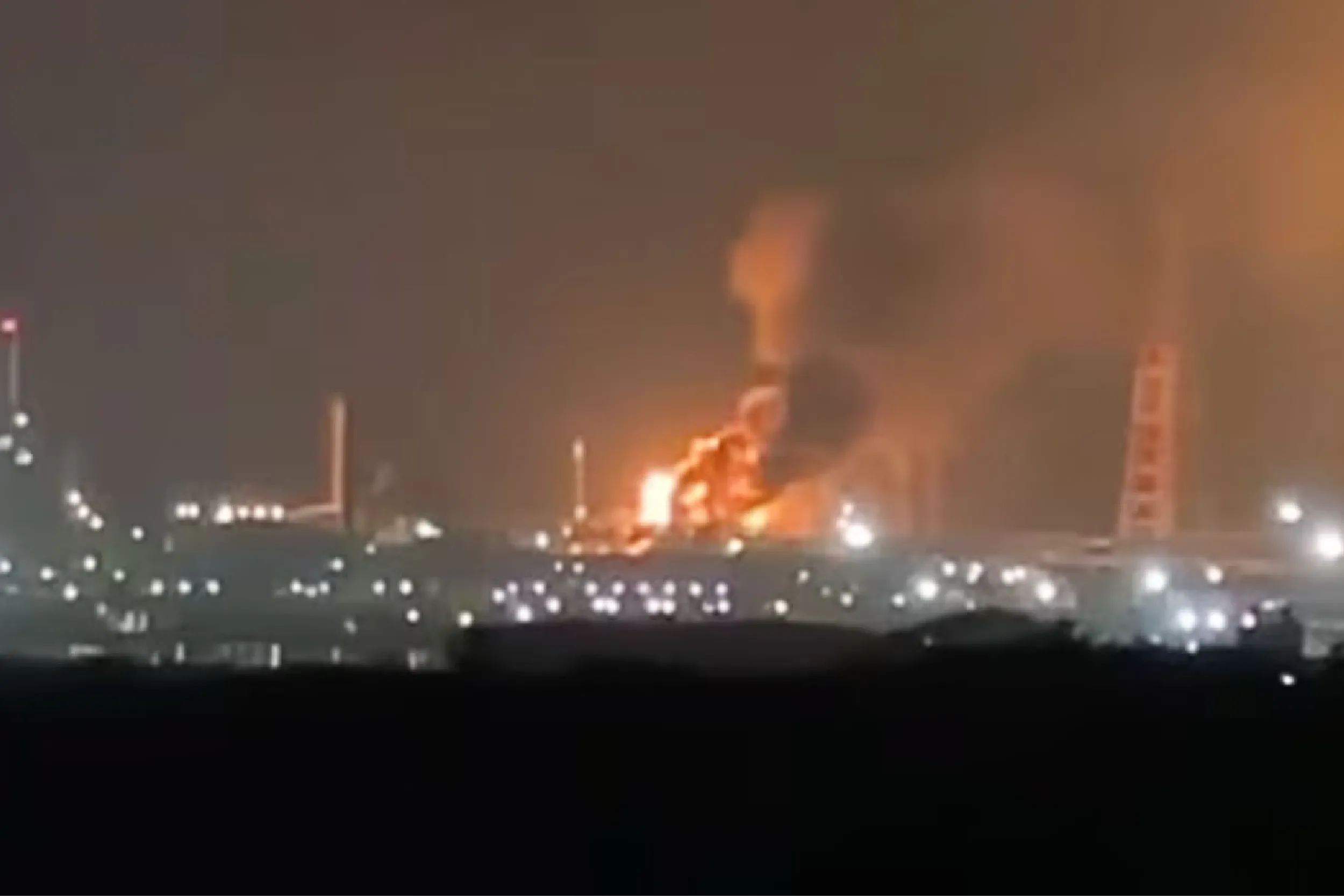 Nighttime cityscape with a large blaze and smoke rising above buildings in the distance; intense orange flames illuminate the scene.