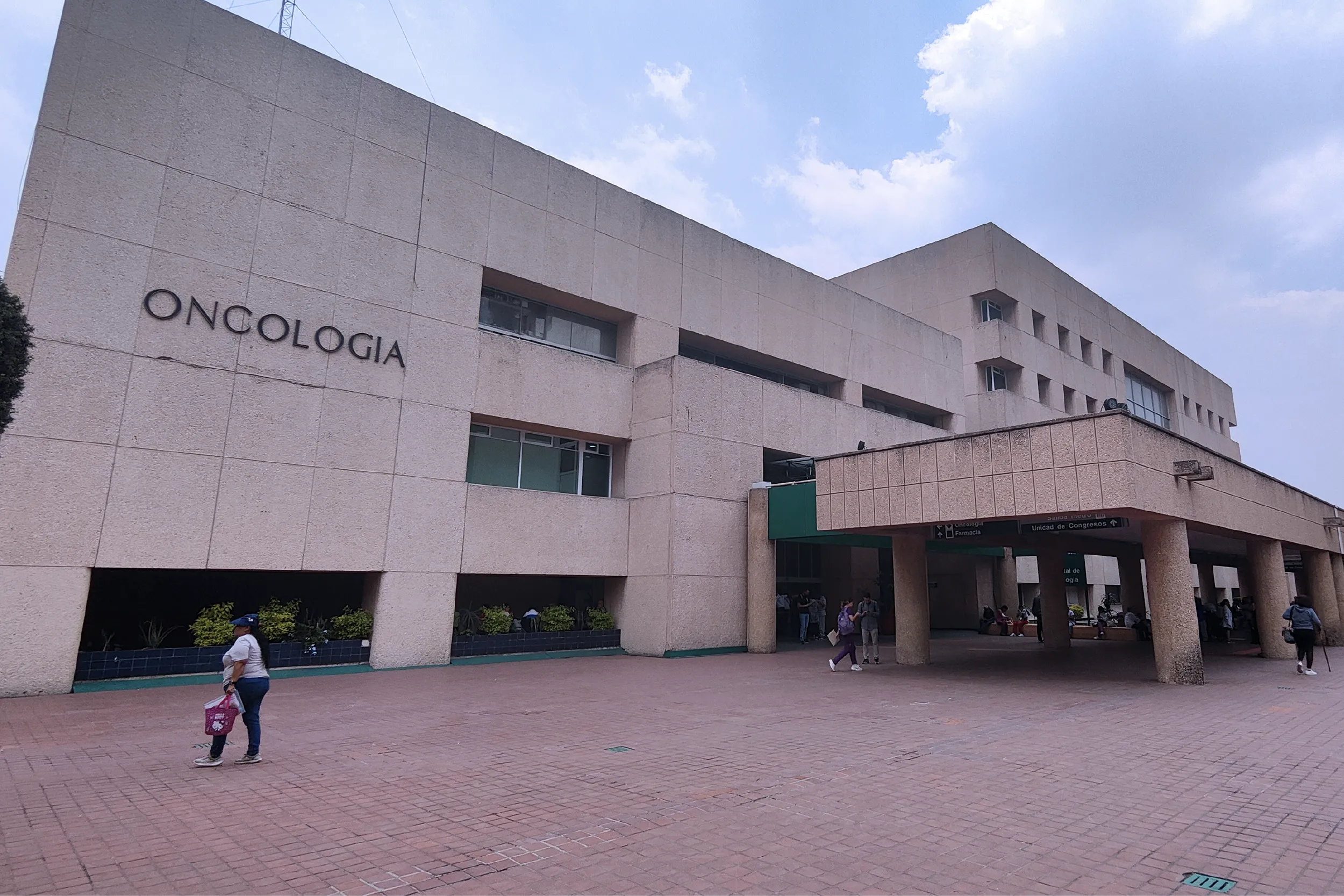 Beige hospital building with the word 'Oncologia' on the wall and people walking in a wide brick plaza under a blue sky.