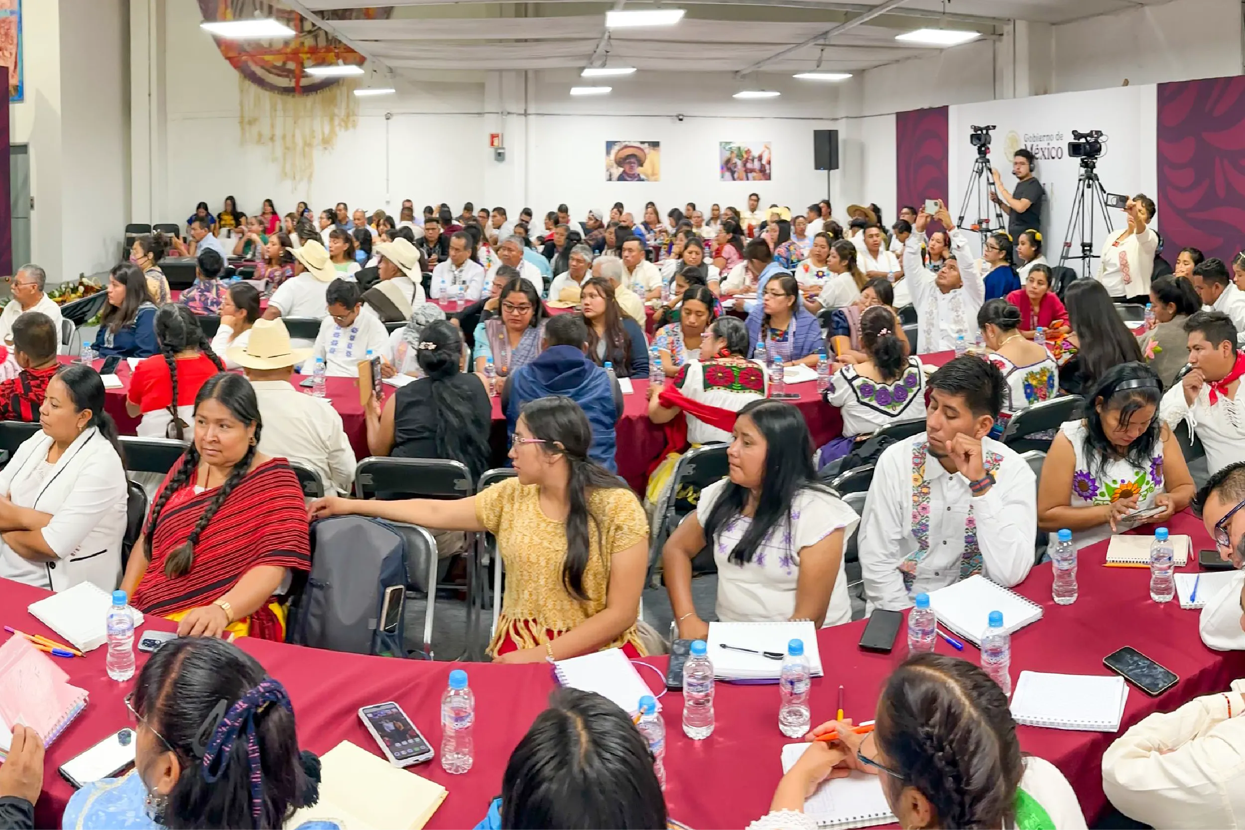 Large hall filled with people seated at red-tablecloth tables for a conference, many in colorful traditional clothing, with cameras on tripods at the back scaling the room.