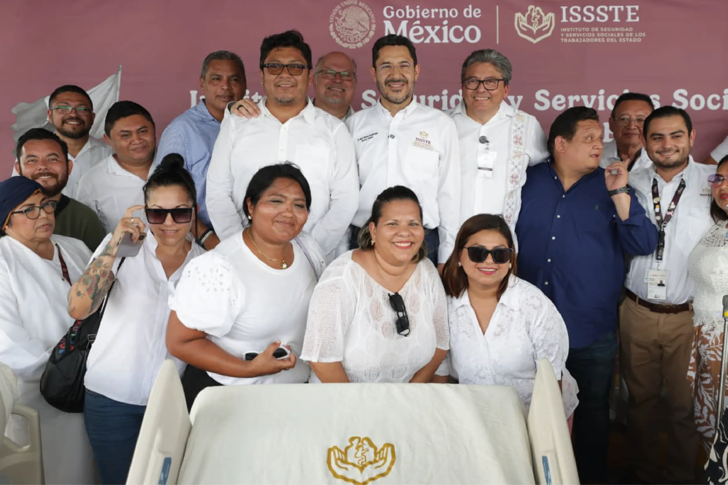 Group of people in white shirts posing for a photo at a formal event, maroon backdrop with Gobierno de México logos behind them