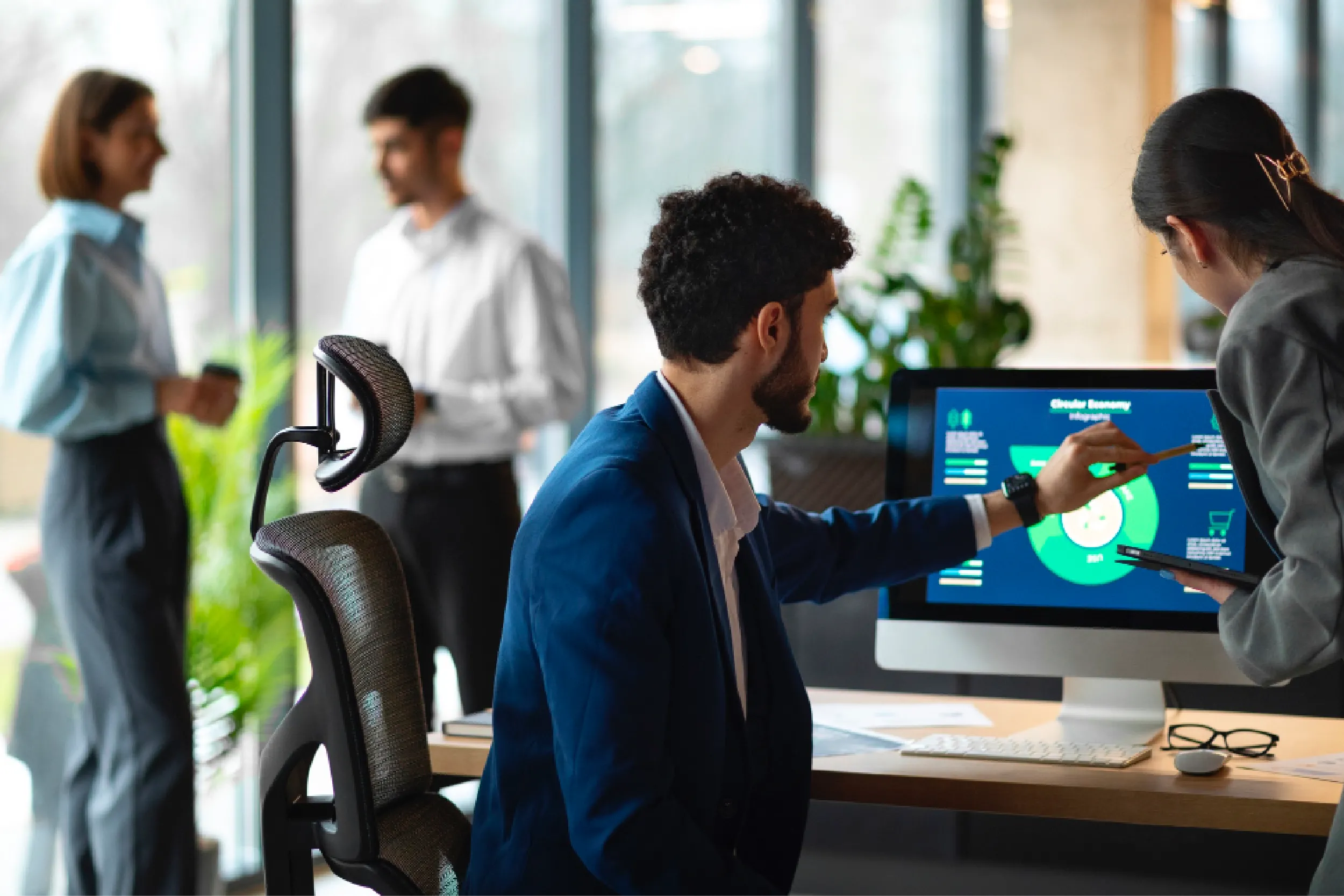 Man in a blazer sits at a desk, pointing with a stylus at a touchscreen monitor displaying charts as a colleague nearby assists. (Office scene)