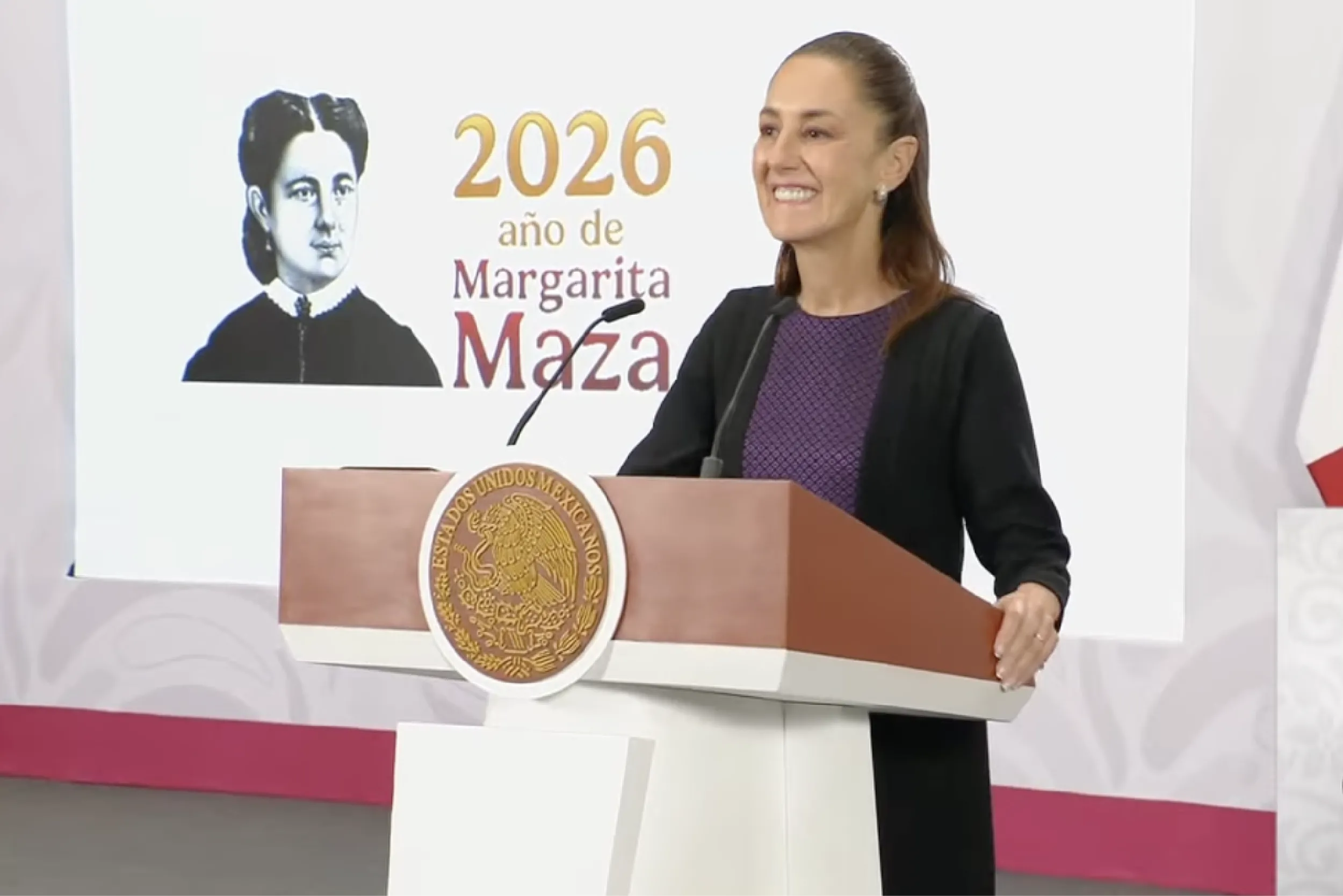 Woman speaker at a podium with a microphone, smiling; backdrop shows '2026 año de Margarita Maz...'; official seal on the podium.