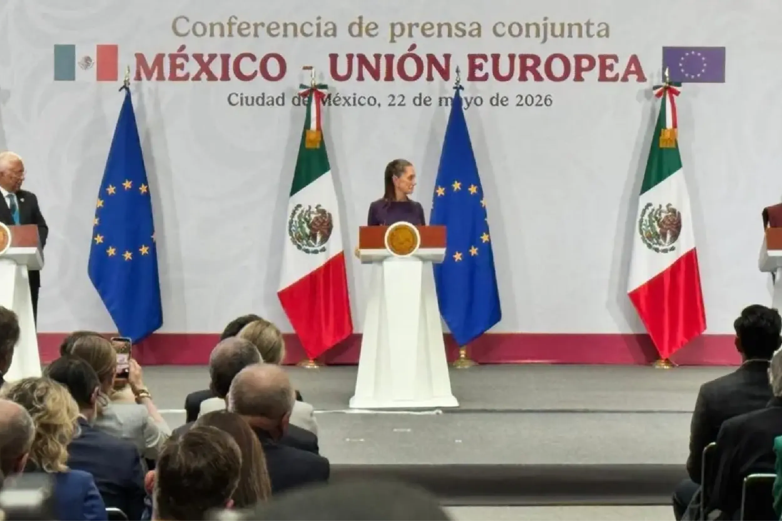 Speaker at a podium during a Mexico–EU joint press conference, with Mexican and European flags on stage and an audience in the foreground.