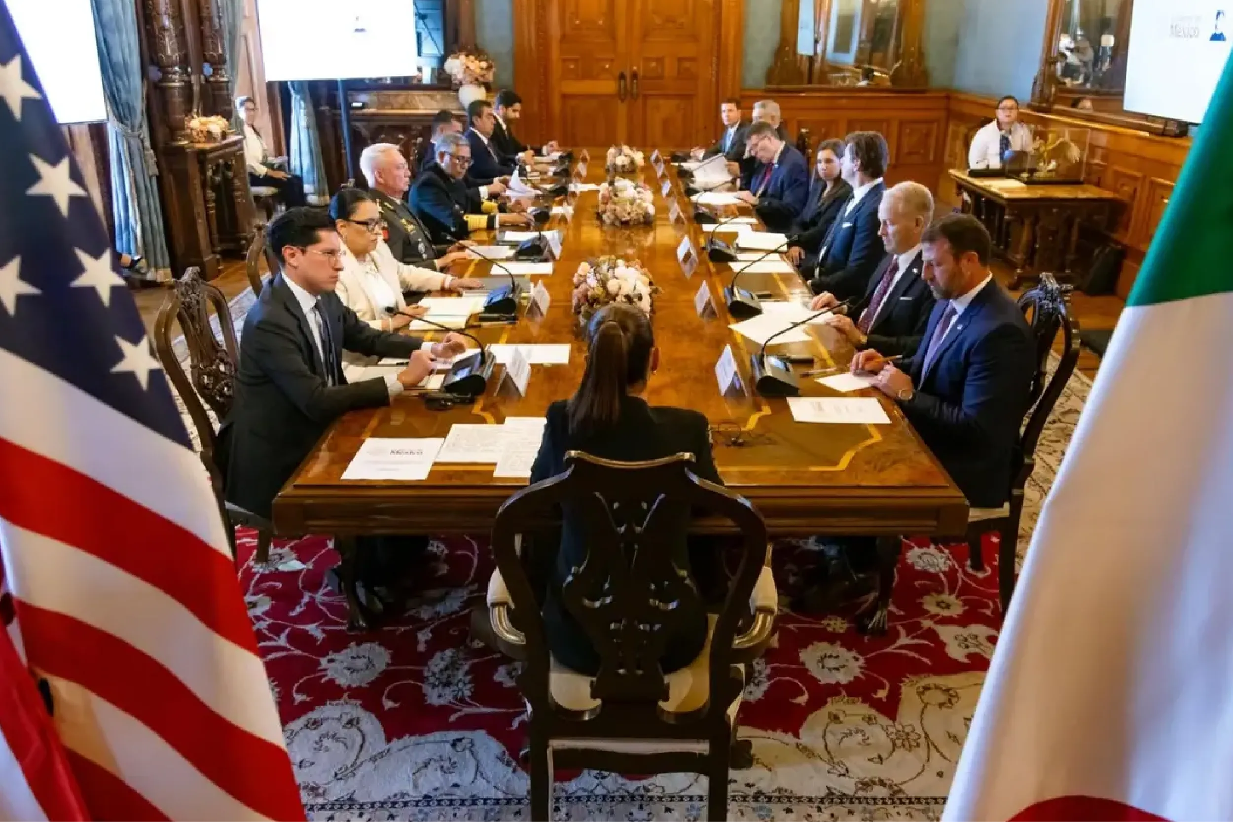 Diplomatic meeting around a long conference table in an ornate wood-paneled room with flags framing the scene.