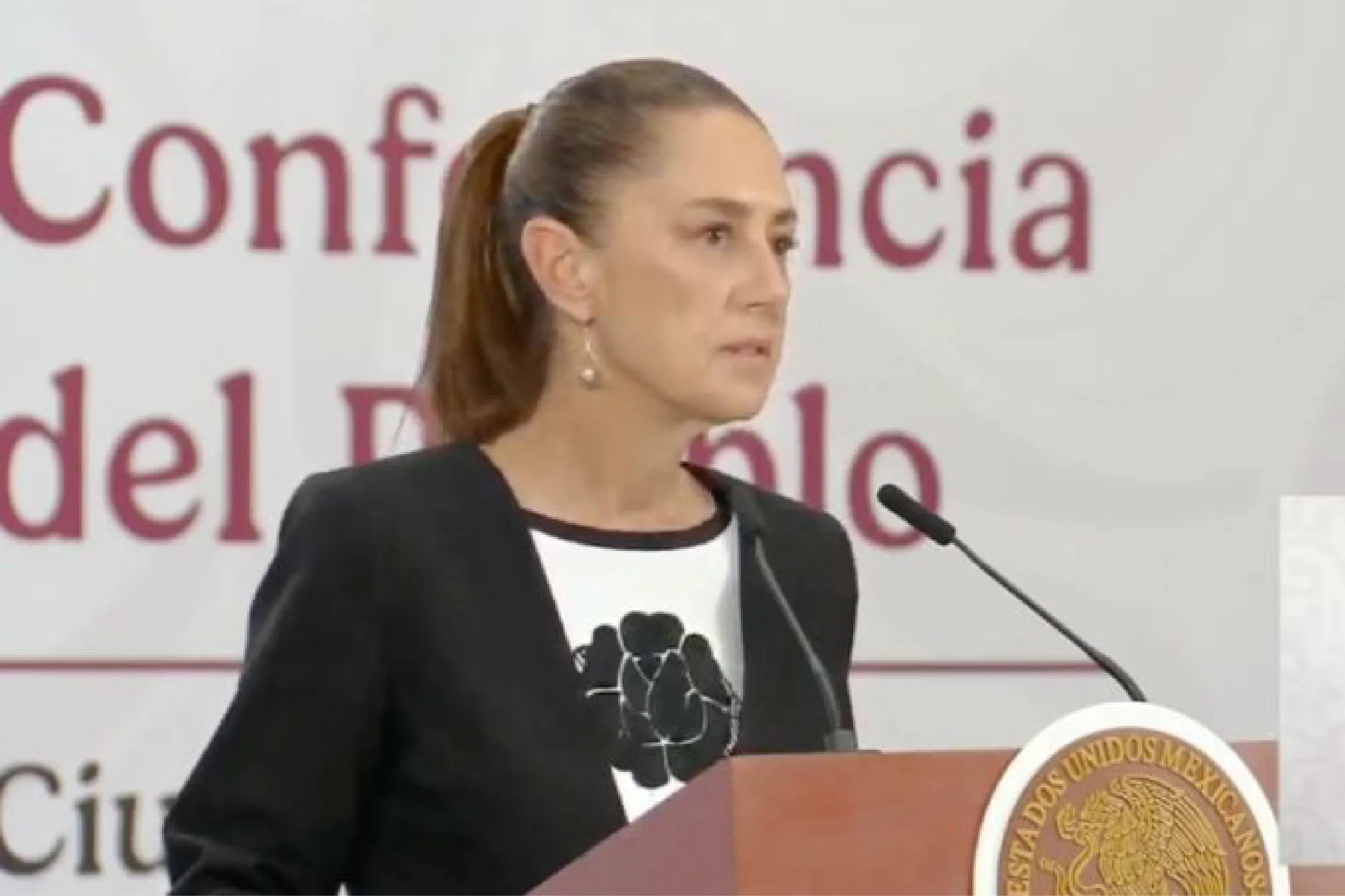 Speaker at a conference podium: woman with ponytail delivering remarks, seal on the lectern, backdrop with large red letters behind her.