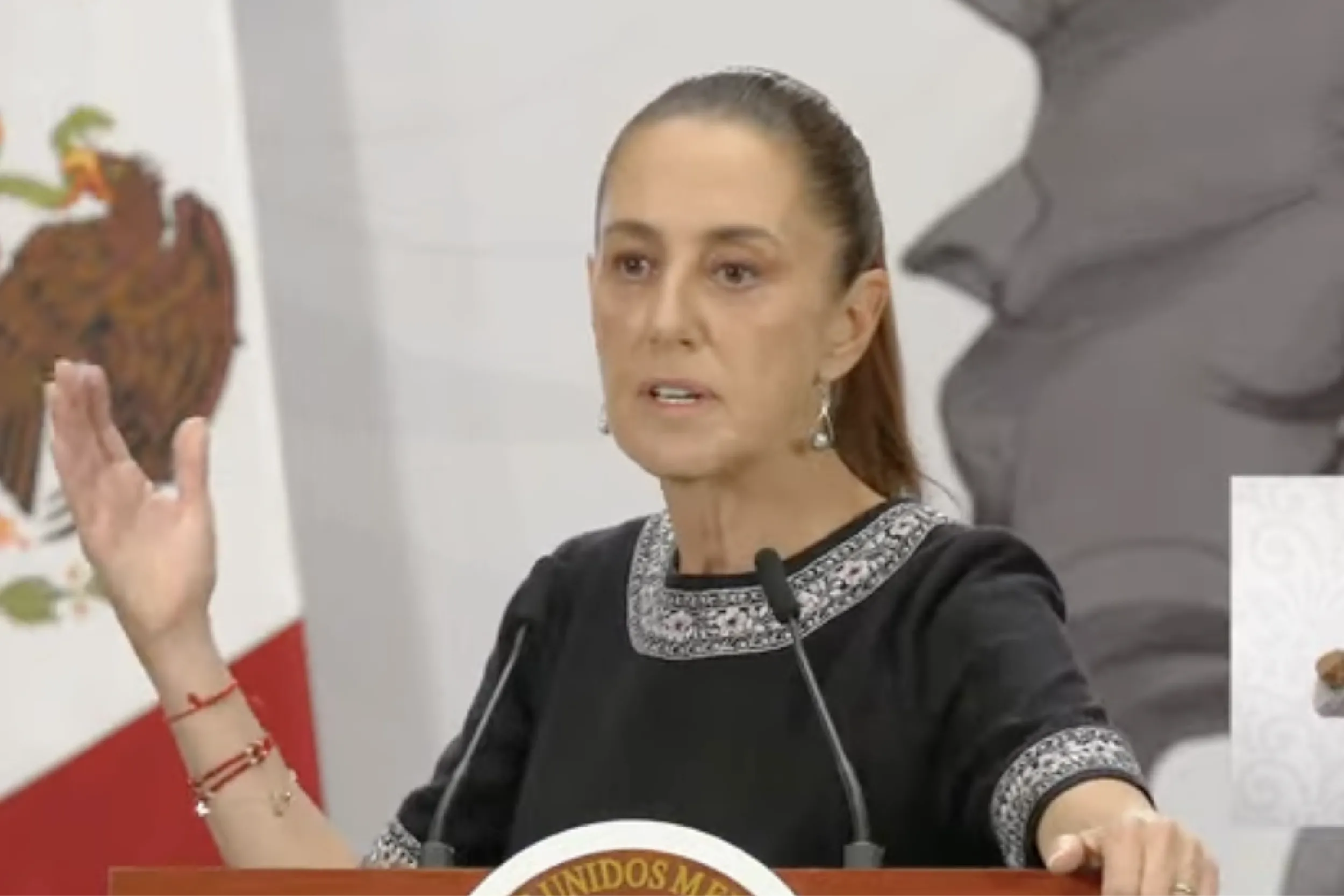 Woman in a black dress at a podium, raising her left hand while speaking into microphones in a formal setting.