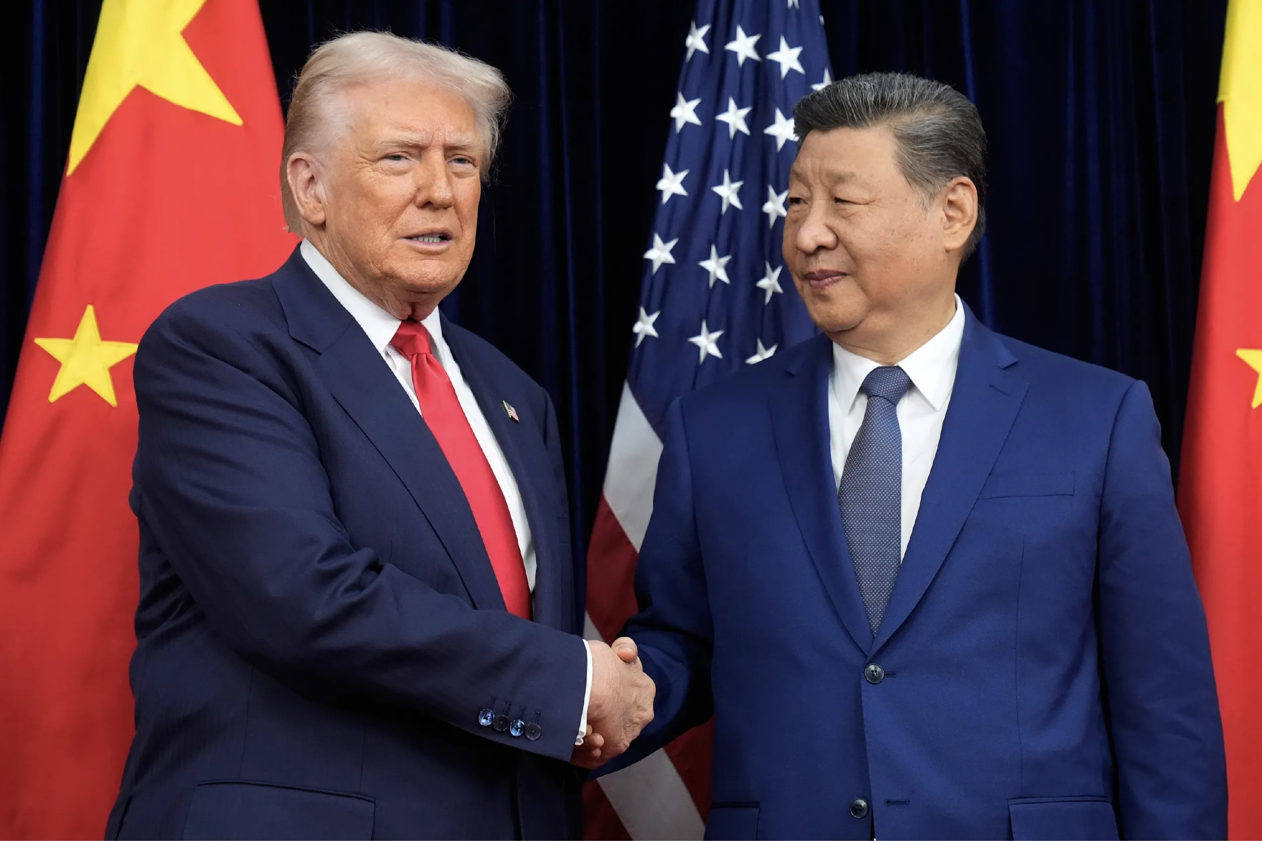 Two men in dark suits shake hands in front of a backdrop of national flags at a formal event.