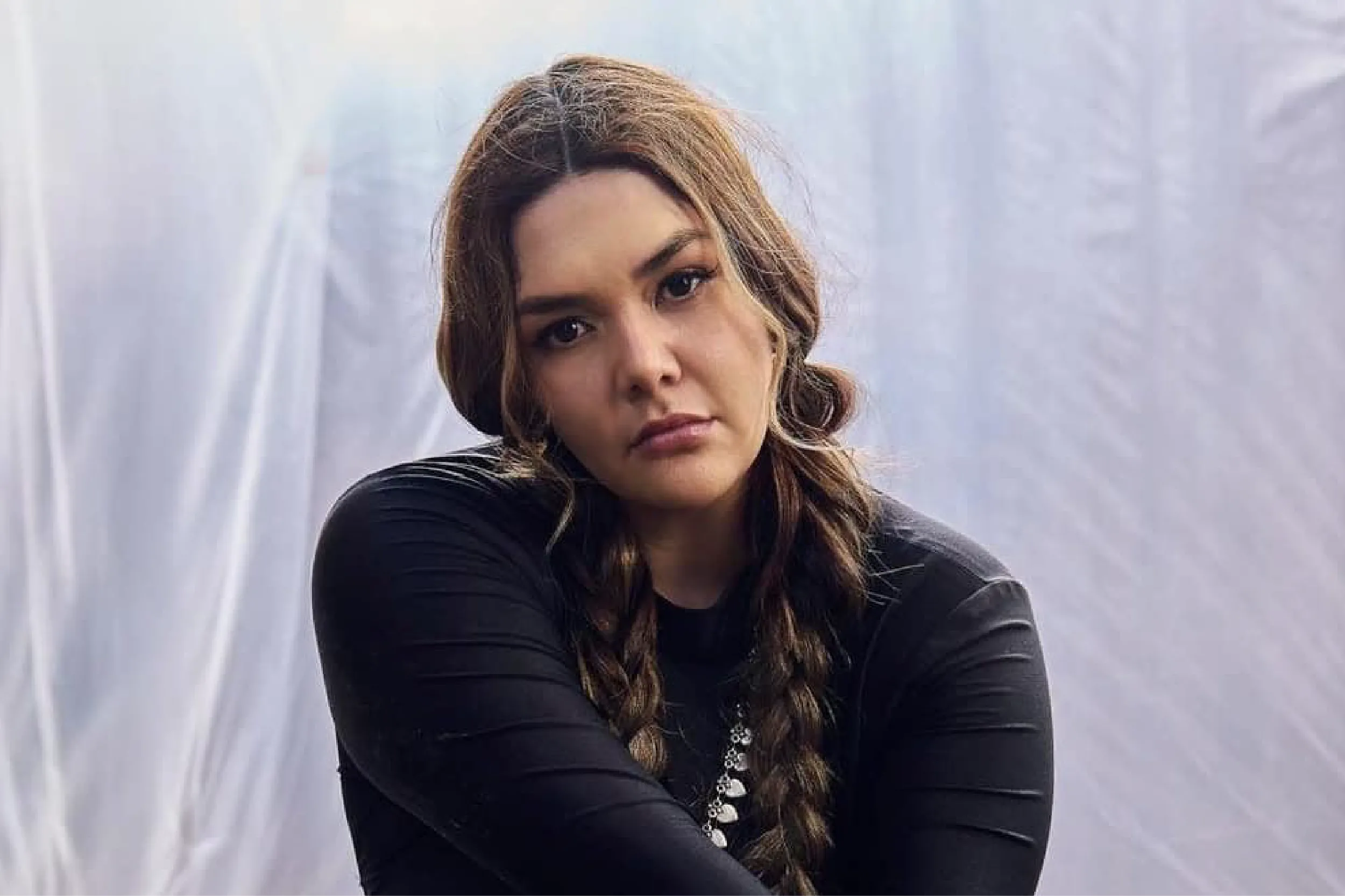 Portrait of a woman with long braided hair wearing a black top, looking at the camera against a pale backdrop.