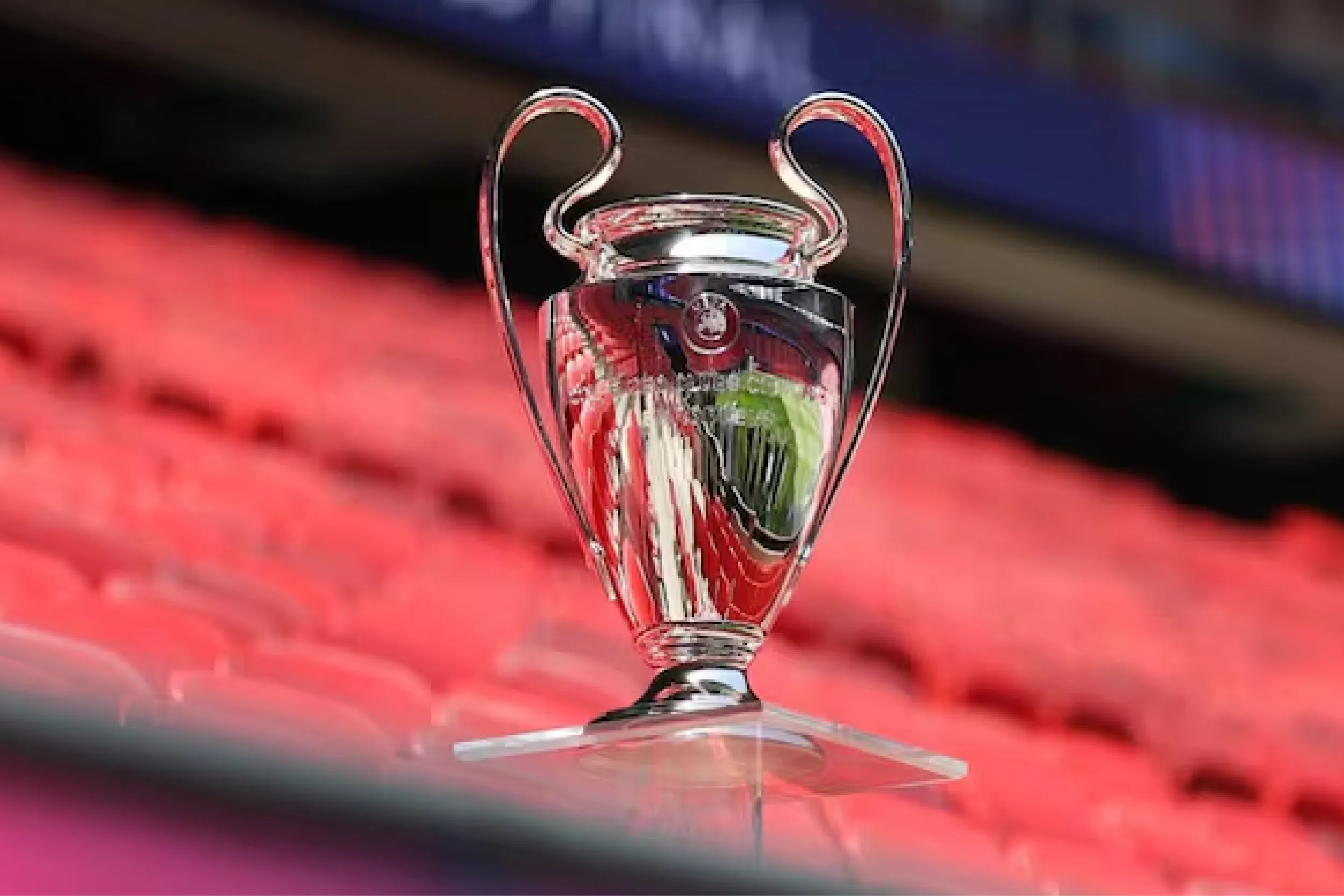 Shiny silver championship trophy on a glass stand with red stadium seating blurred in the background.