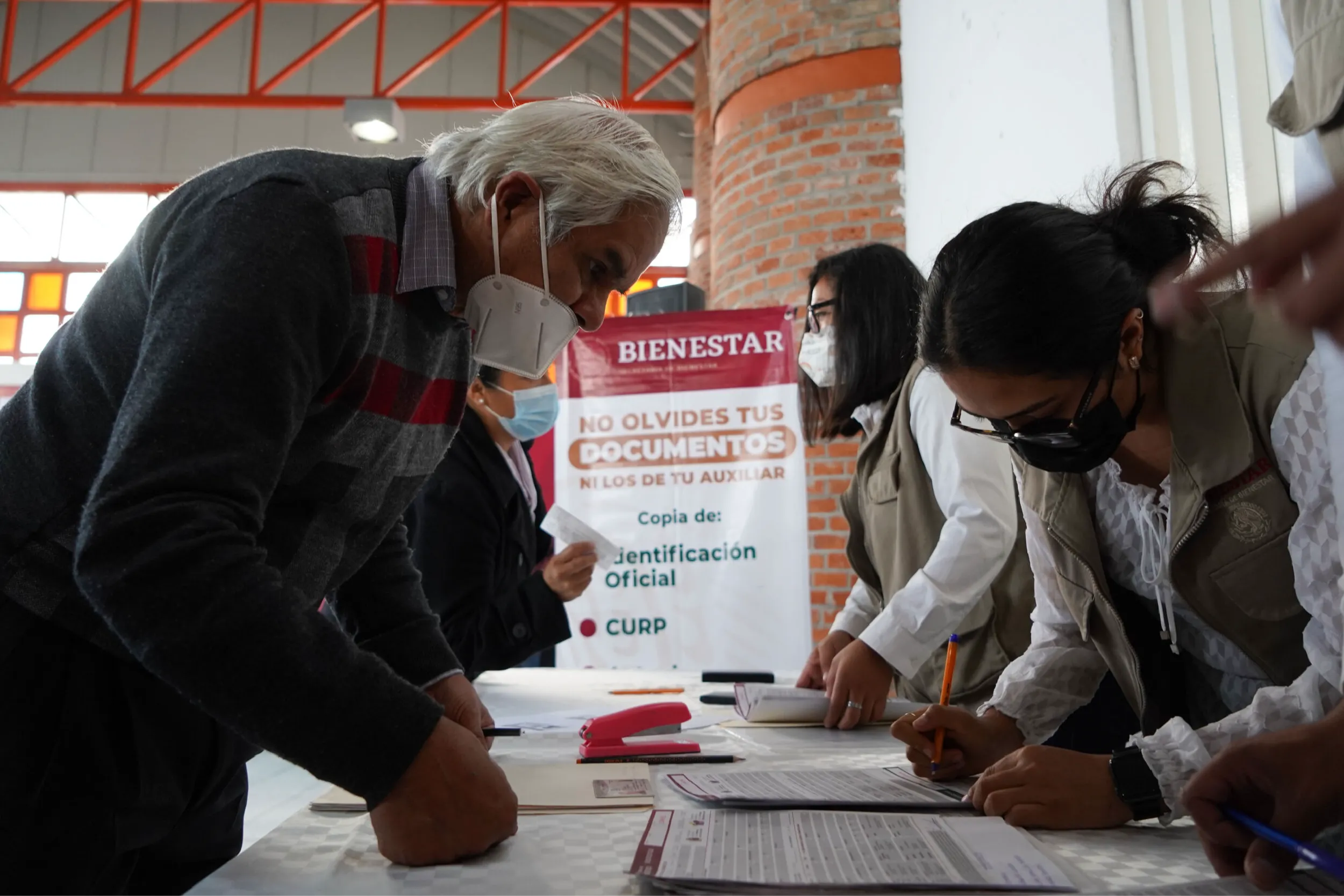 People wearing masks lean over a table, filling out forms as staff assist beside a red banner that reads No olvides tus documentos (Do not forget your documents).