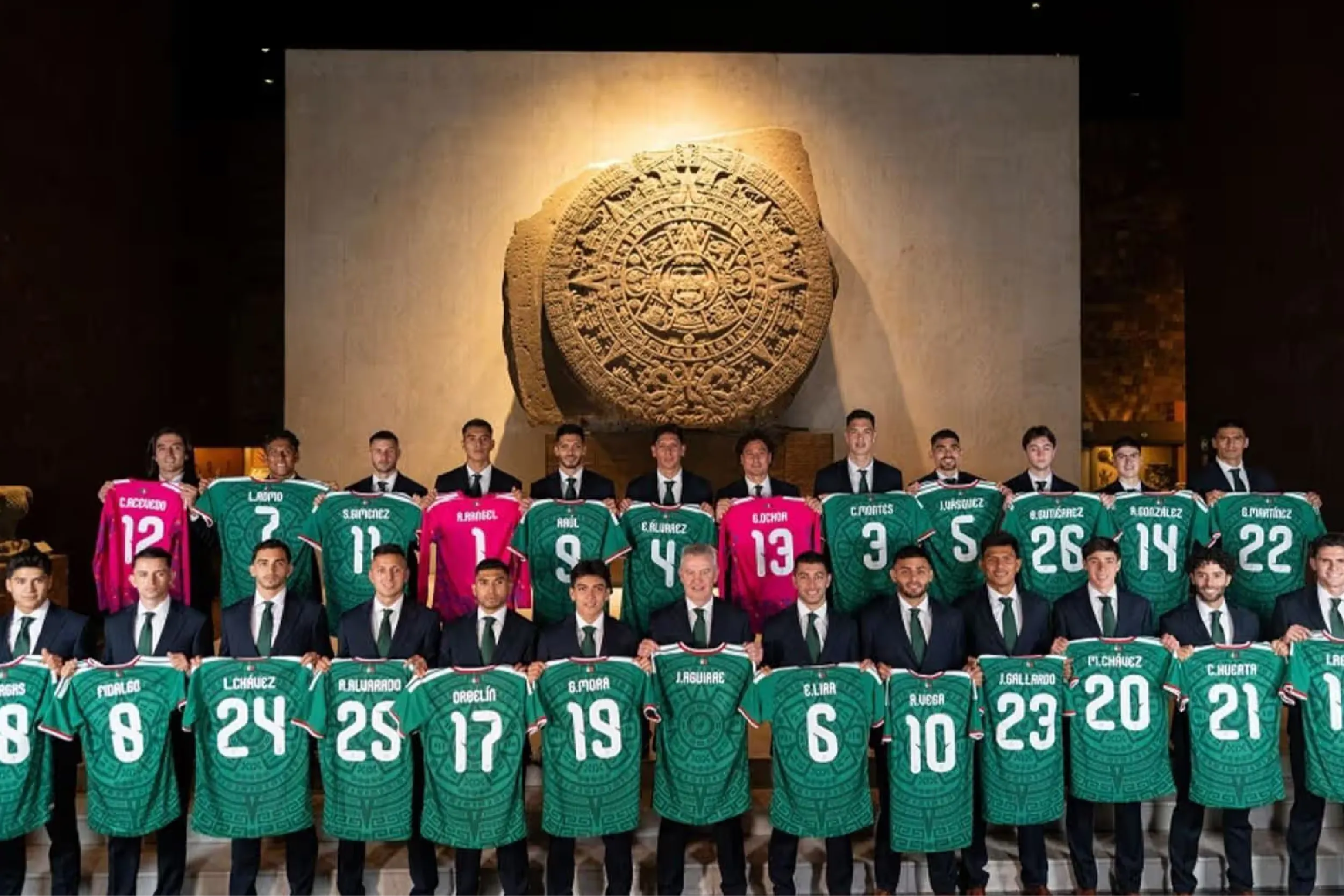Group of soccer players in suits holding green and pink jerseys, posed in front of a large stone Aztec calendar artifact in a museum backdrop.