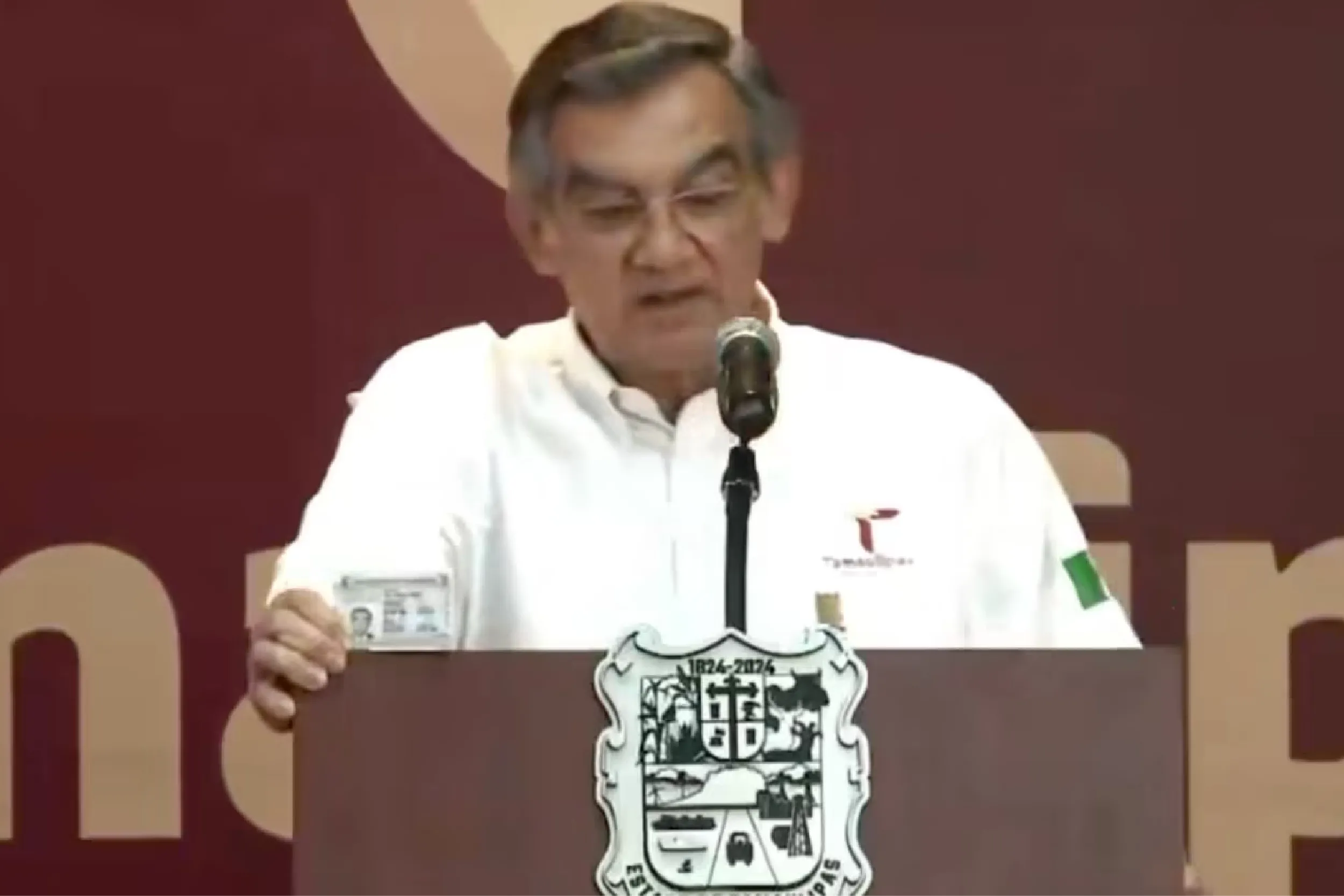 Man in a white shirt speaking at a podium with a microphone during a formal event, badge visible on chest area.