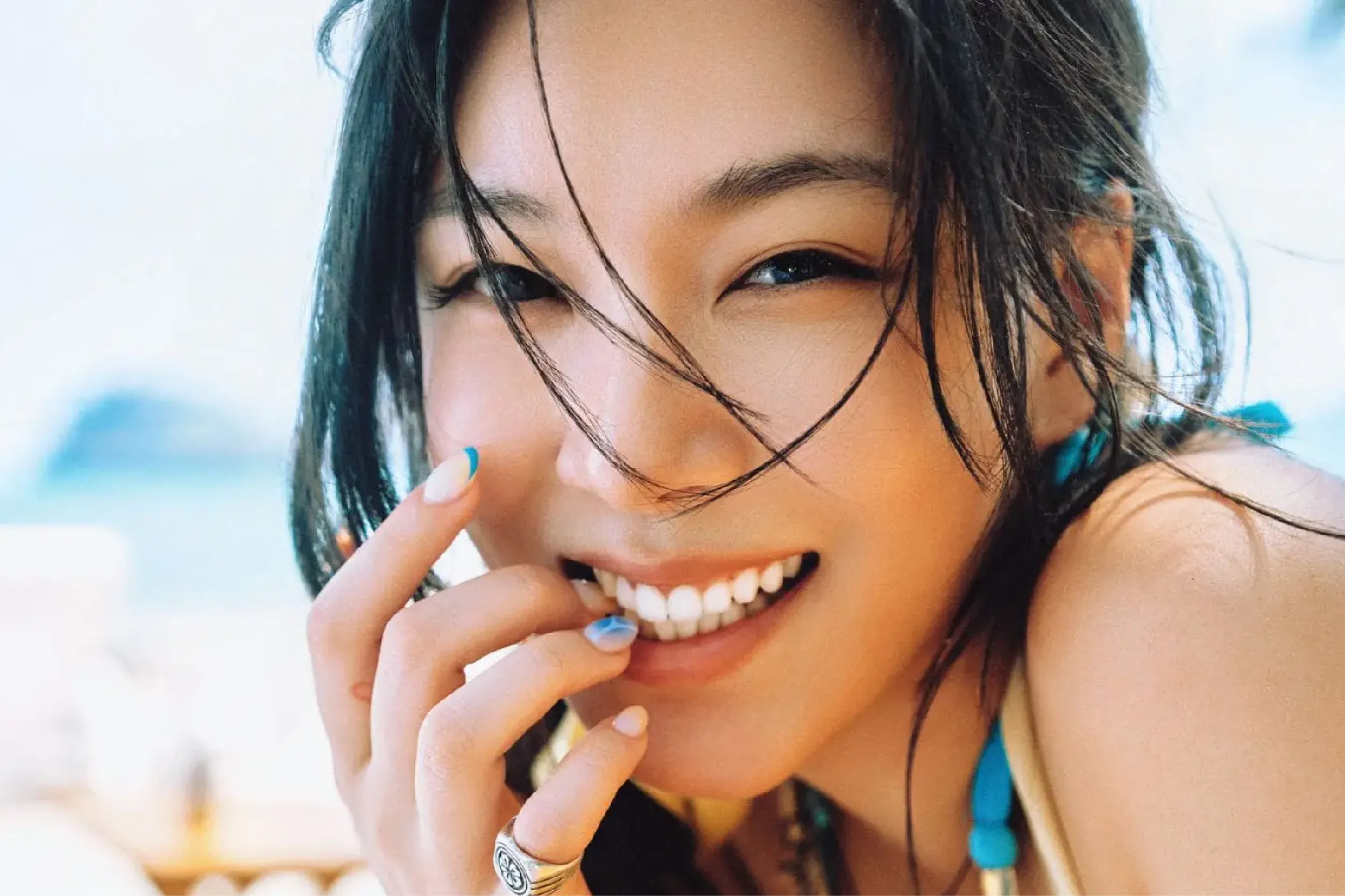 Close-up of a smiling woman with wet hair, touching her lips with a blue manicure and turquoise bead accessories, outdoors on a sunny day