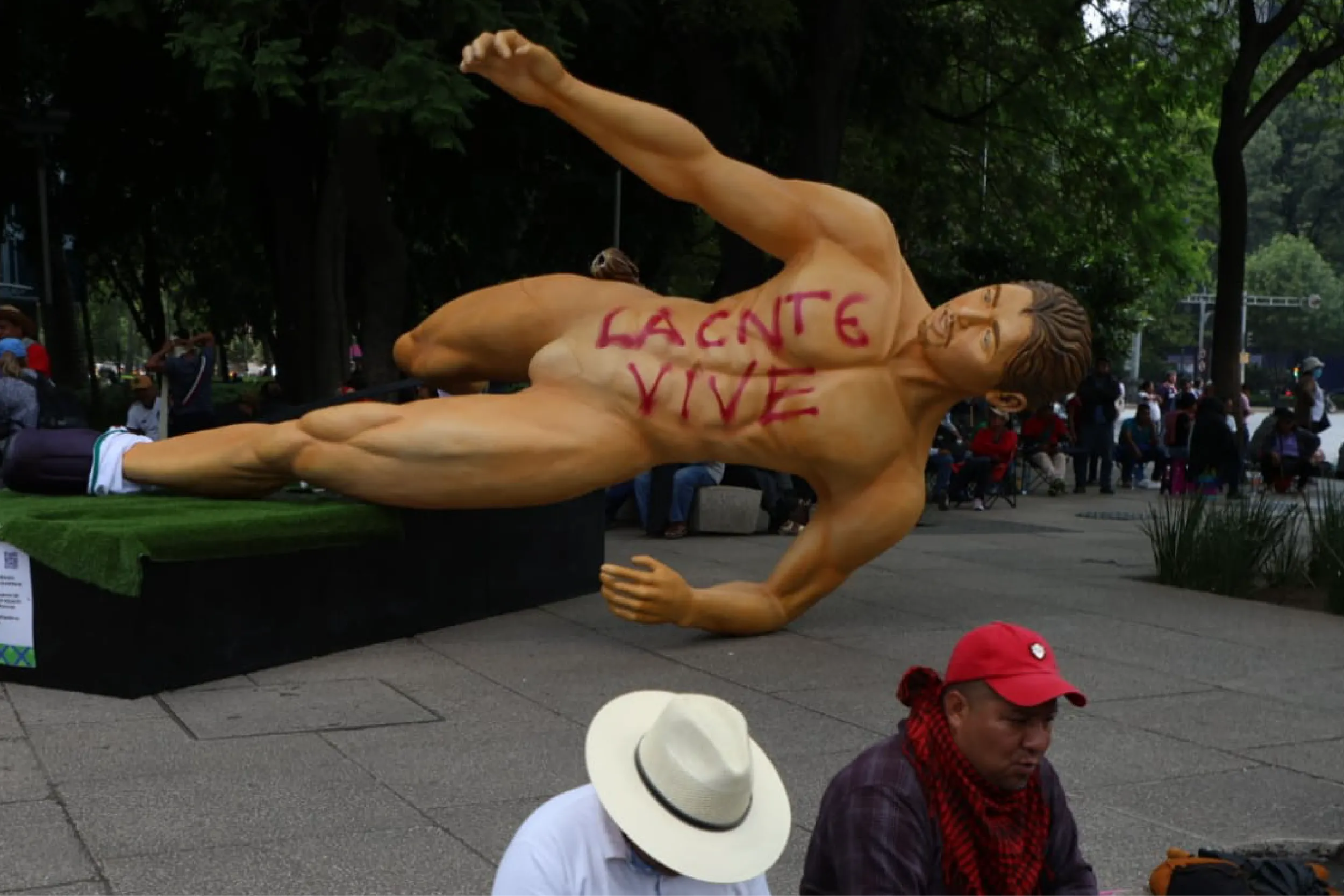 Large reclining nude statue with red graffiti-like text 'LACNTE VIVE' on chest, displayed in a park with onlookers nearby.
