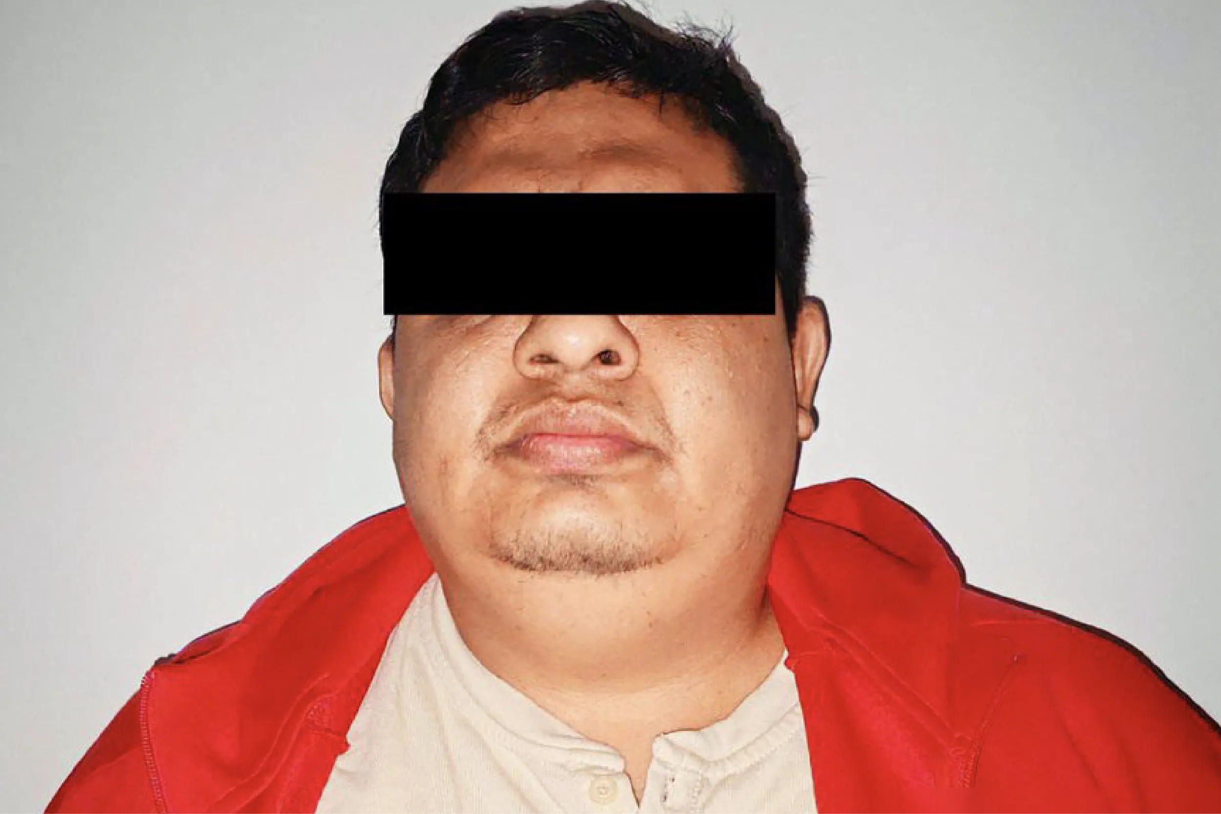 Portrait of a man facing the camera with his eyes covered by a black censor bar; wearing a red jacket over a white shirt against a neutral background.