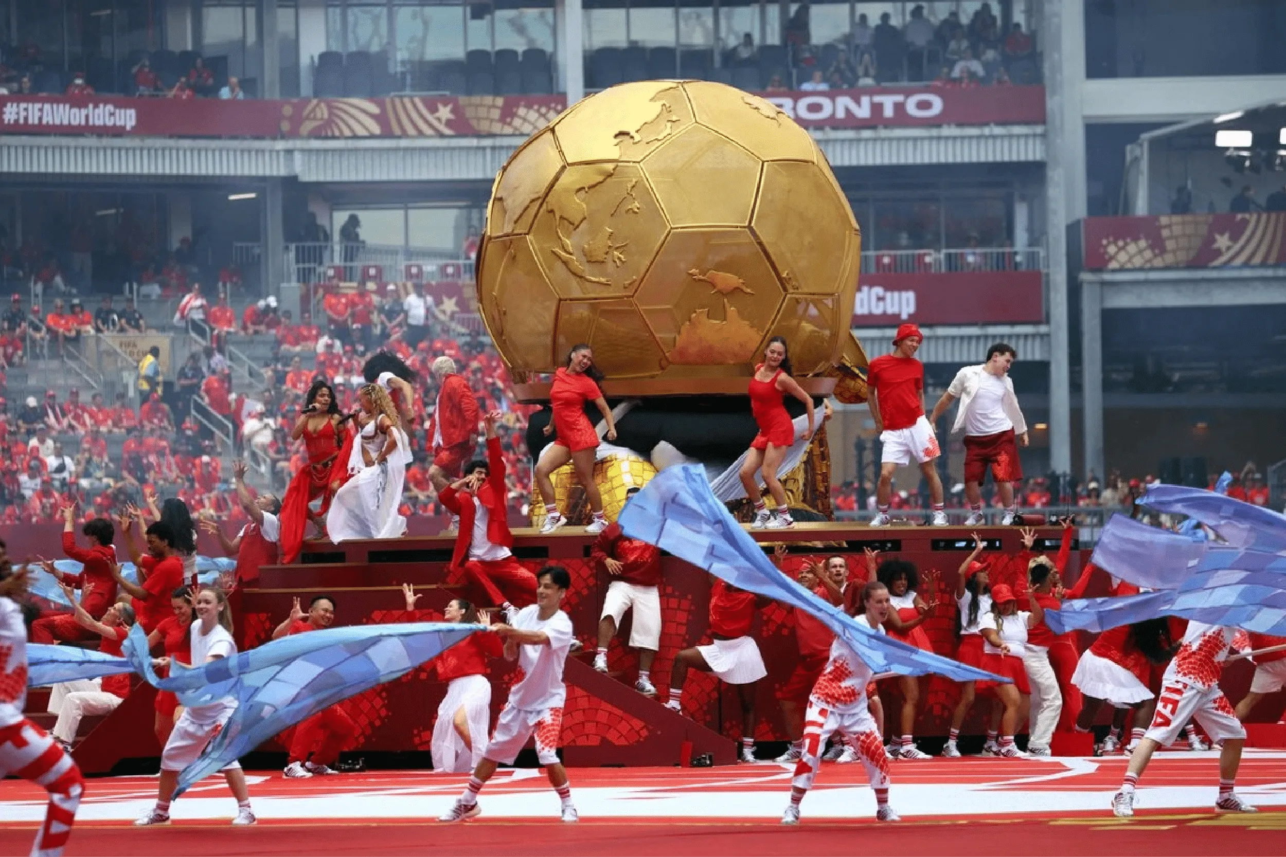 Giant golden spherical World Cup prop on a parade float, with dancers in red and white and flowing blue scarves in a stadium crowd background.