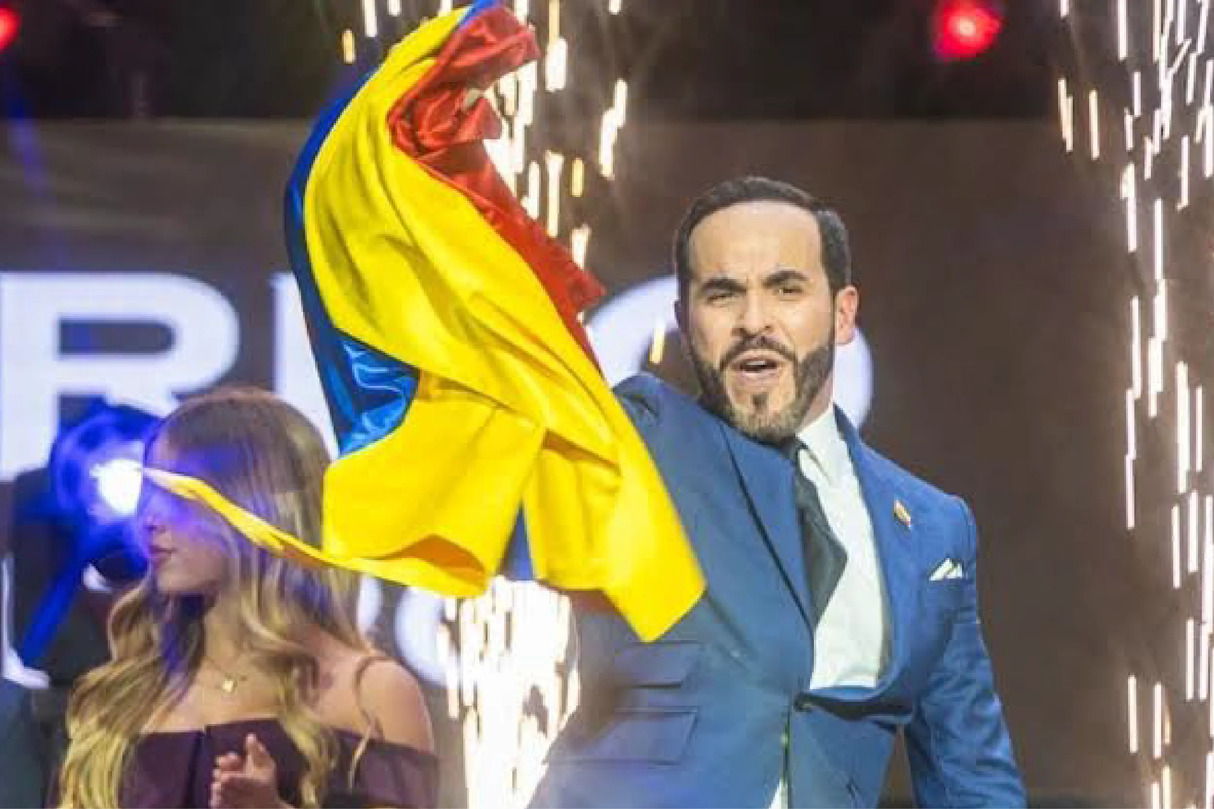 Man in a blue suit waves a Colombian flag on stage amid fireworks and cheering crowd at a celebration.