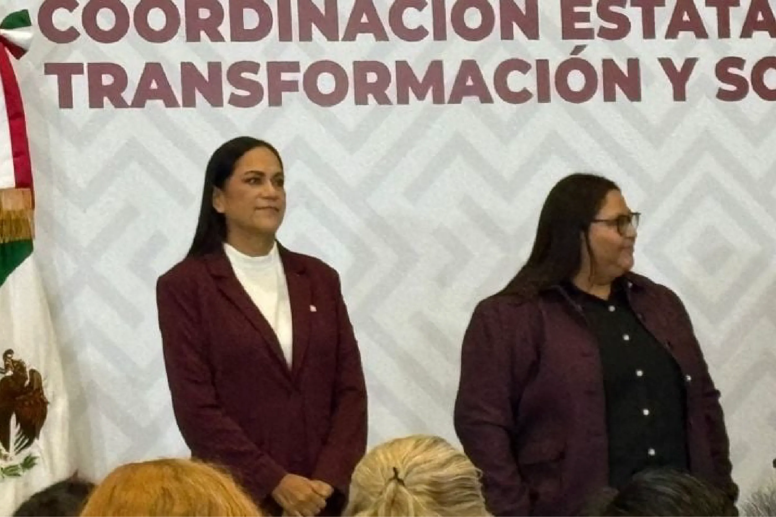 Two women in burgundy blazers stand on a stage in front of a banner with Spanish text; a flag is visible on the left.