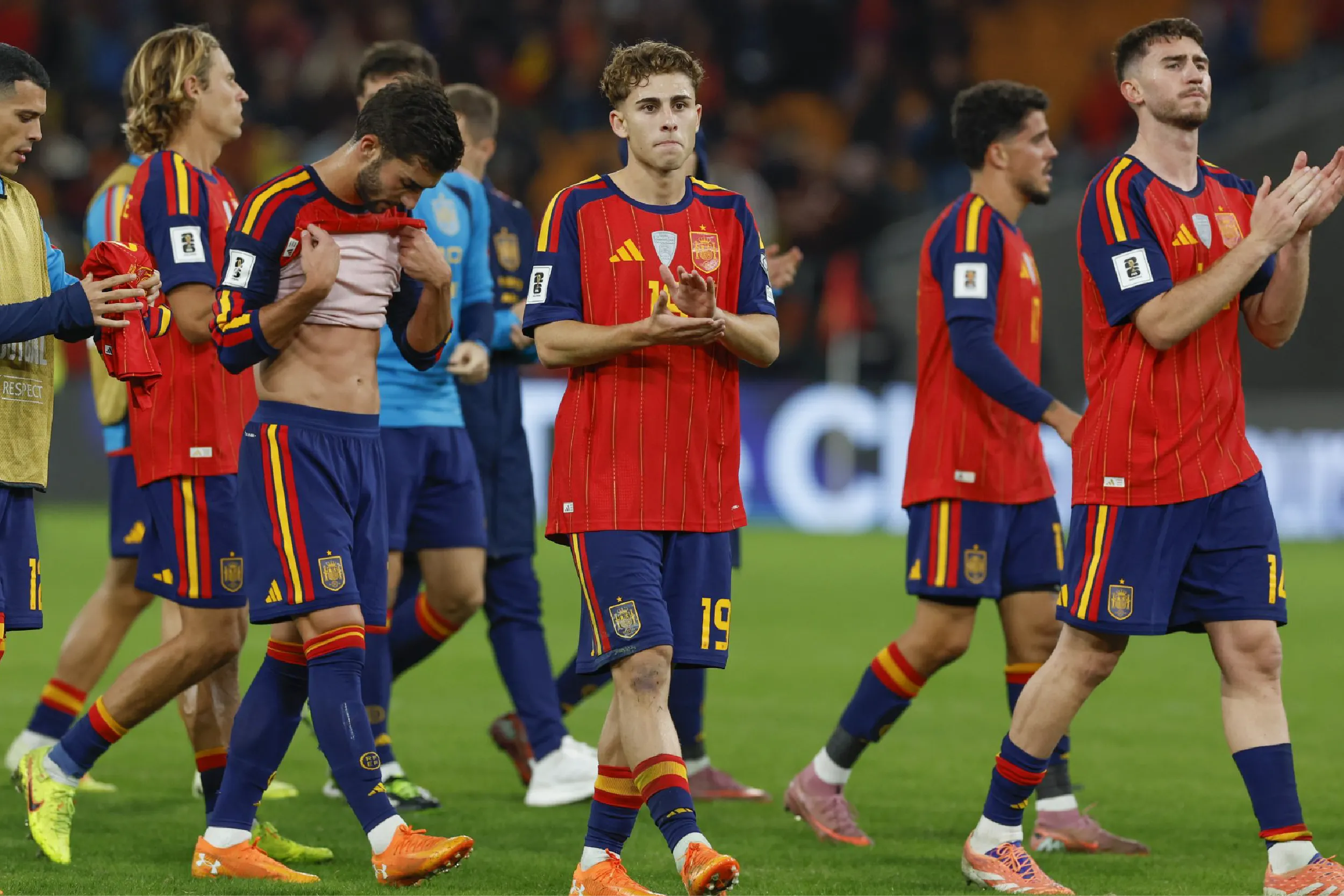 Group of Spain soccer players in red and blue kits walk on a green pitch, some clapping as they leave the field.
