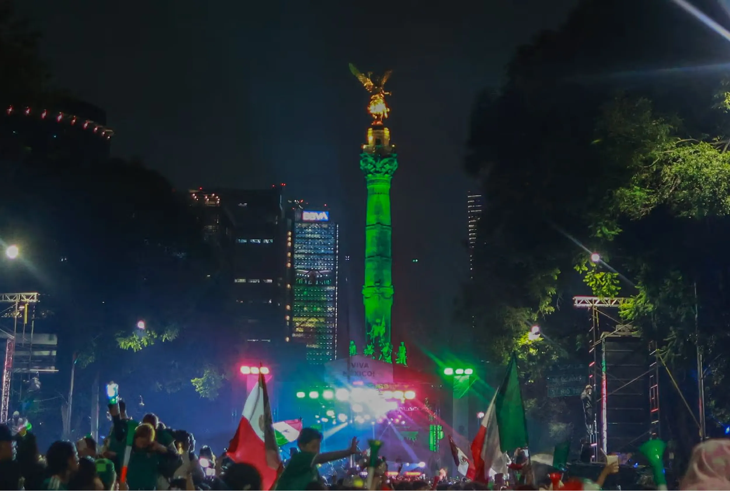 Night view of a city square with a tall monument lit in green and gold at night, surrounded by protesters waving Mexican flags.
