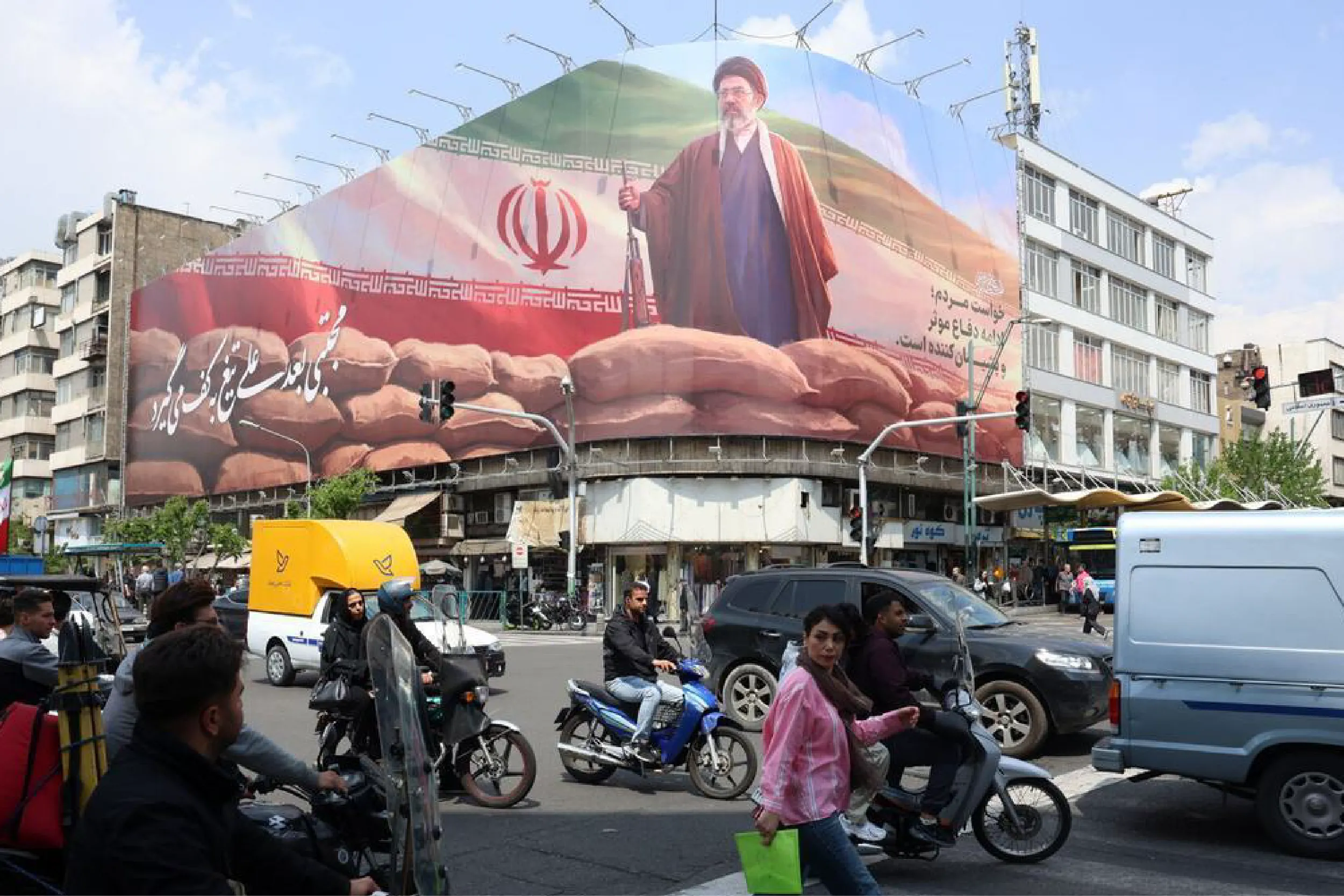 Busy city intersection with motorcycles and pedestrians; a giant mural of Ayatollah Khomeini on a building.