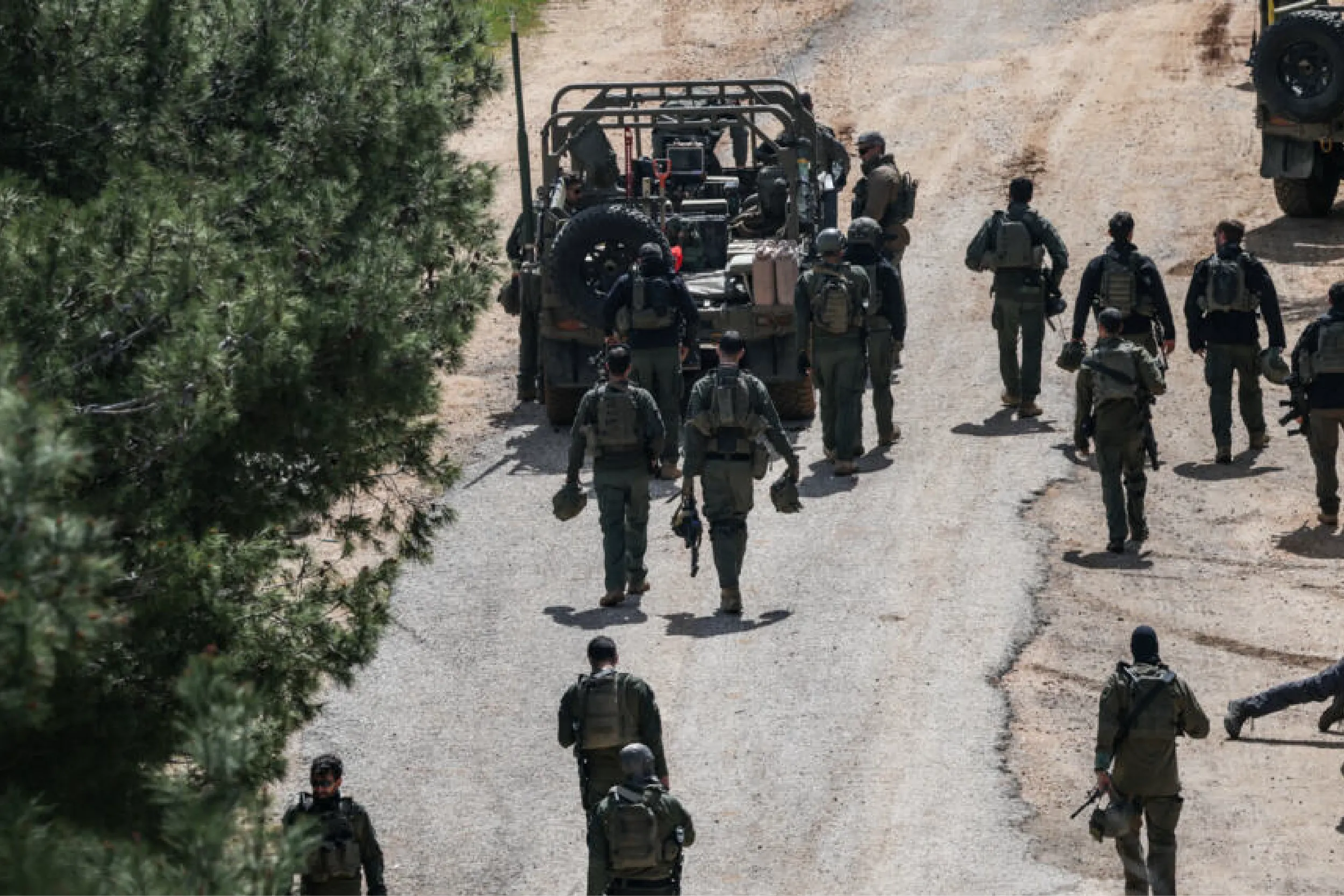 Group of soldiers in tactical gear gathered around a military transport vehicle on a dirt road.