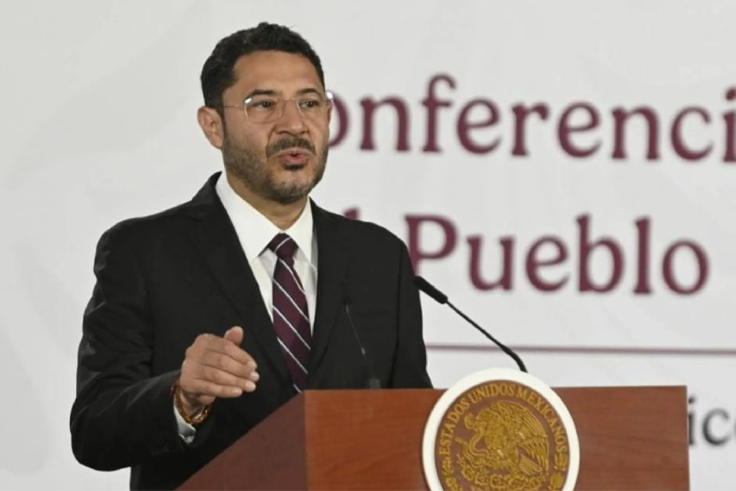 A man in a dark suit and striped tie speaks at a podium with a gold seal, gesturing with his hand.