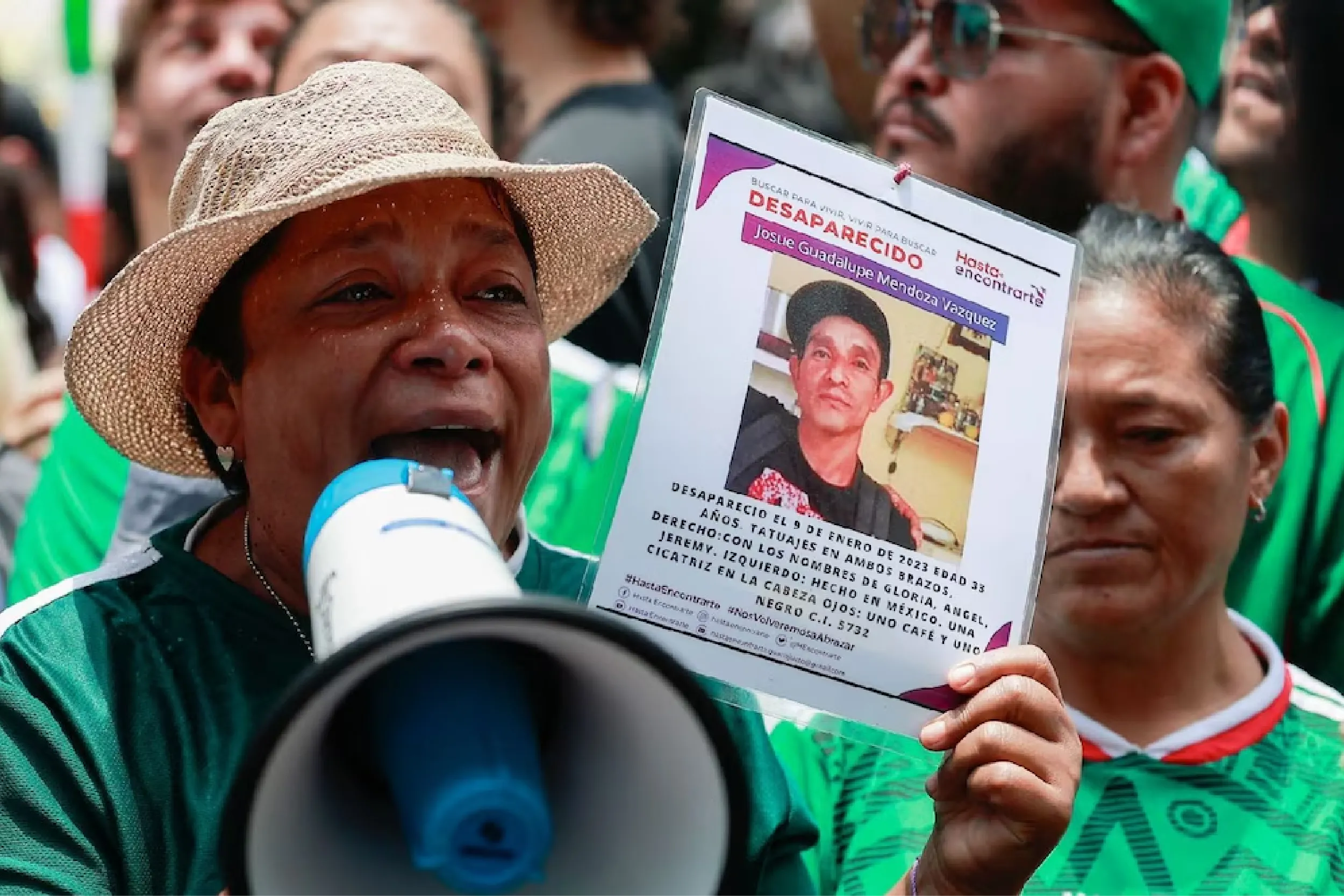 A woman wearing a straw hat shouts into a megaphone while holding a flyer about a missing person at a protest, with others in green shirts in the background.