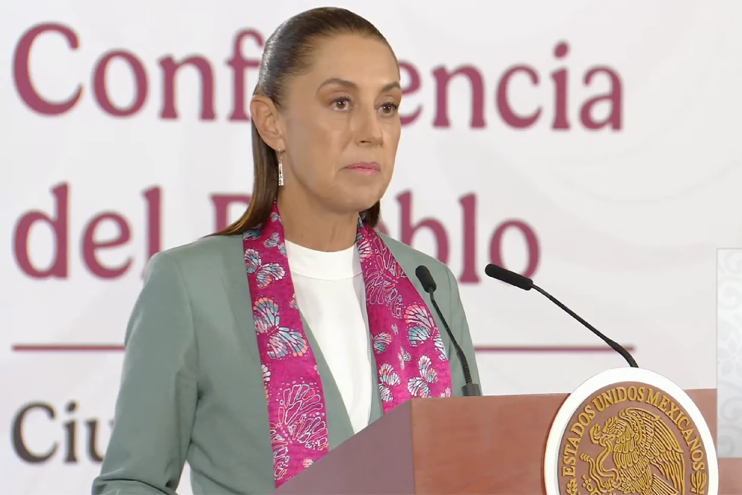 Woman at a podium delivering a conference speech, with microphones and a large official seal visible on the podium behind her