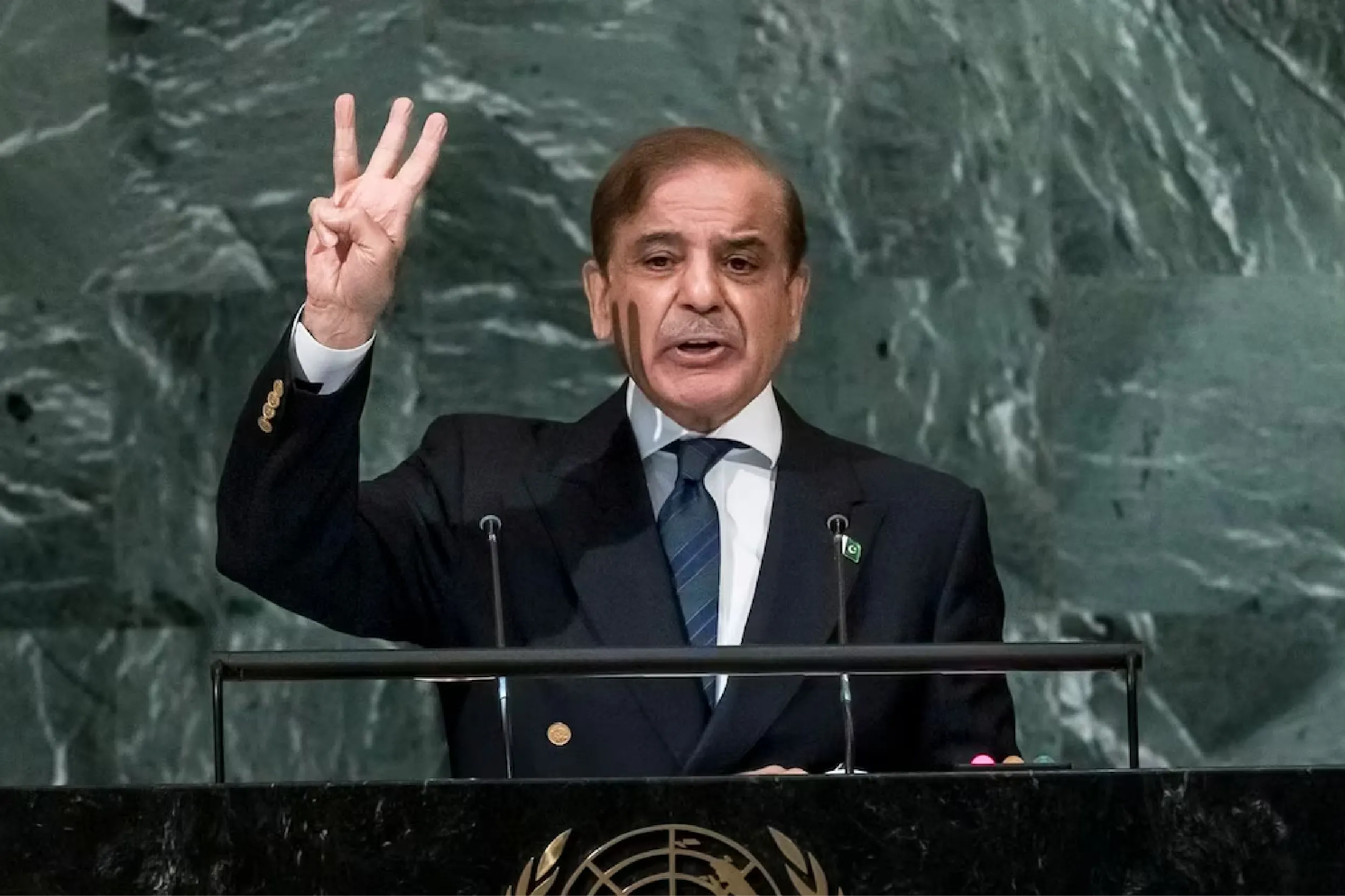 Male speaker at a United Nations podium, raising three fingers in a gesture, wearing a dark suit and tie in front of a marble backdrop.