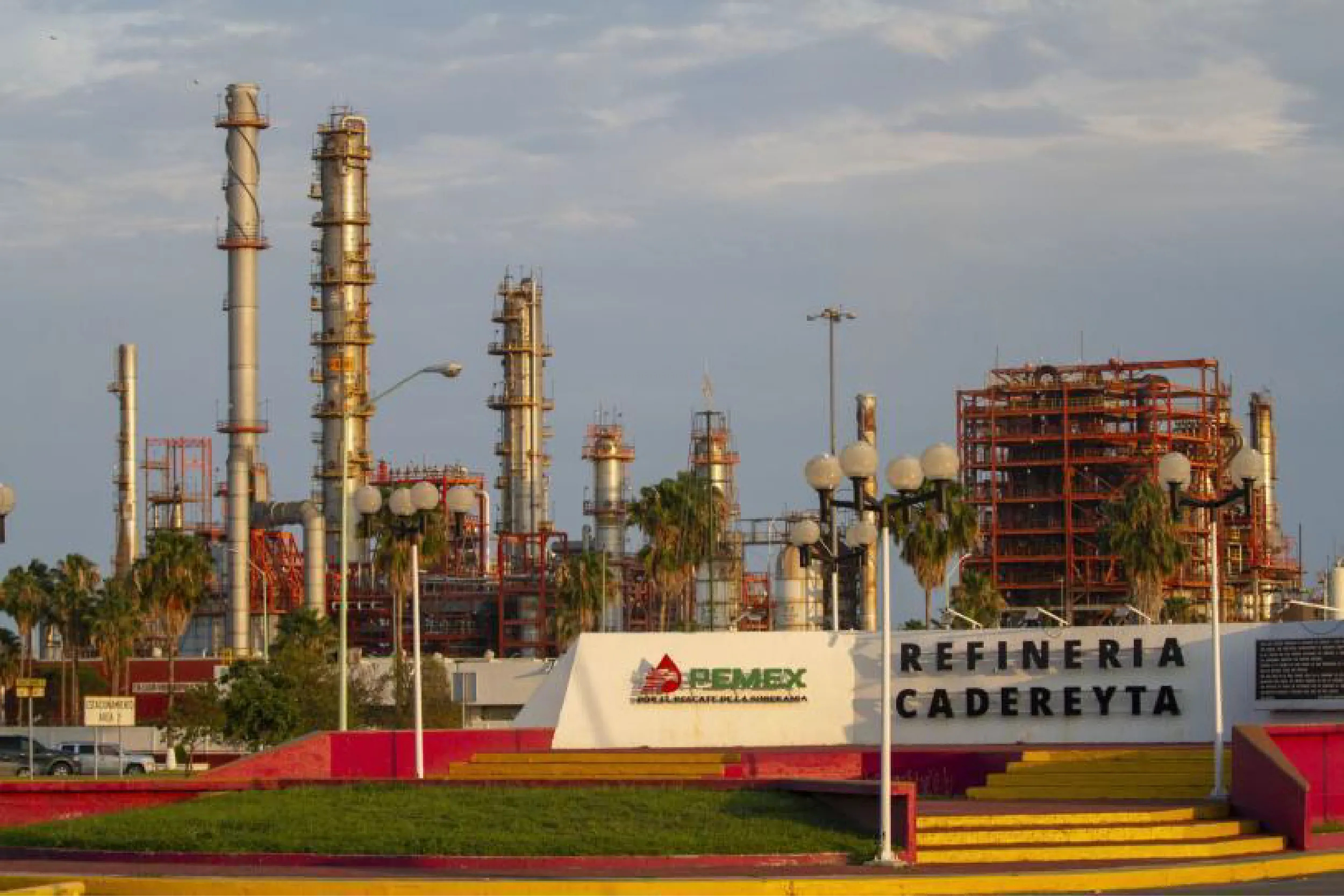 Industrial refinery scene with tall distillation towers, orange-red piping, and storage structures against a blue sky atop a landscaped foreground.