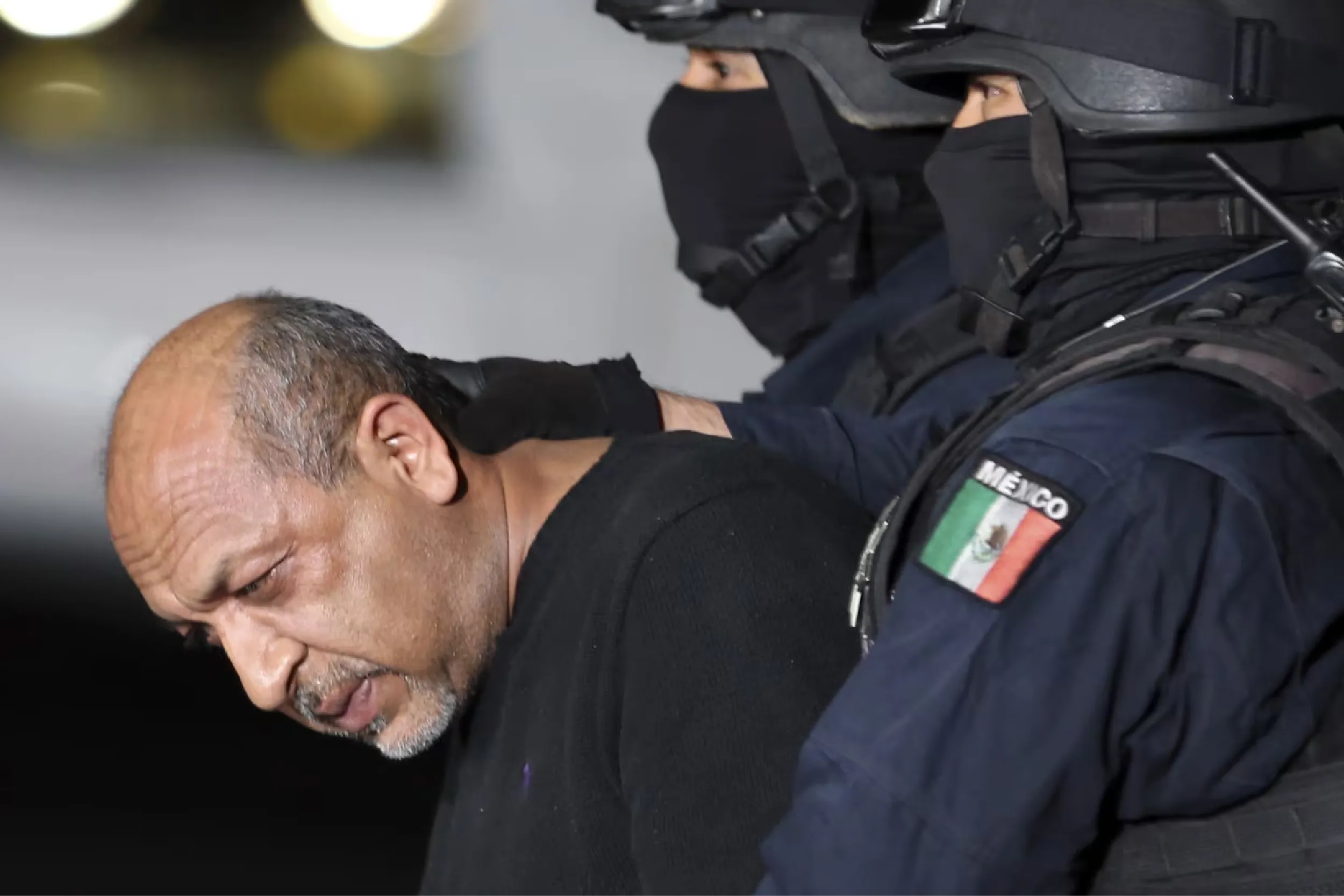 Masked tactical officer in black gear restrains a man, sleeve shows a Mexican flag patch.